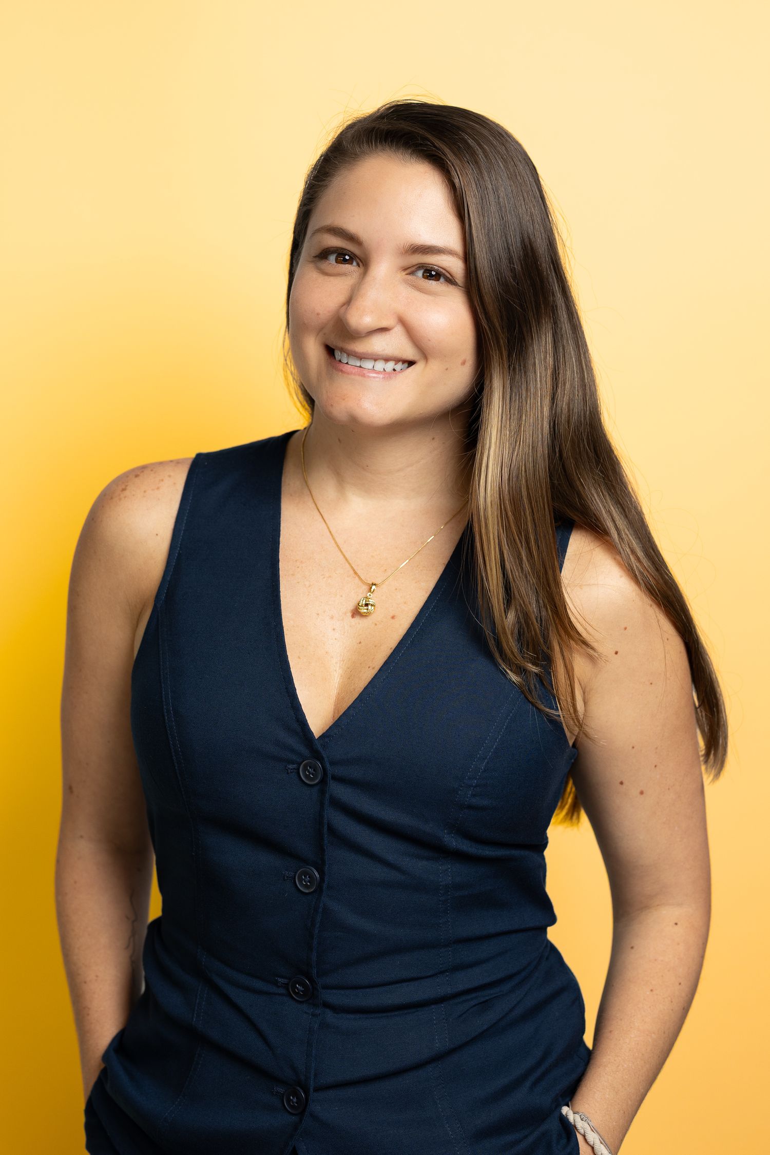 Smiling young woman with long brown hair wearing a navy sleeveless vest and gold necklace against a yellow background.