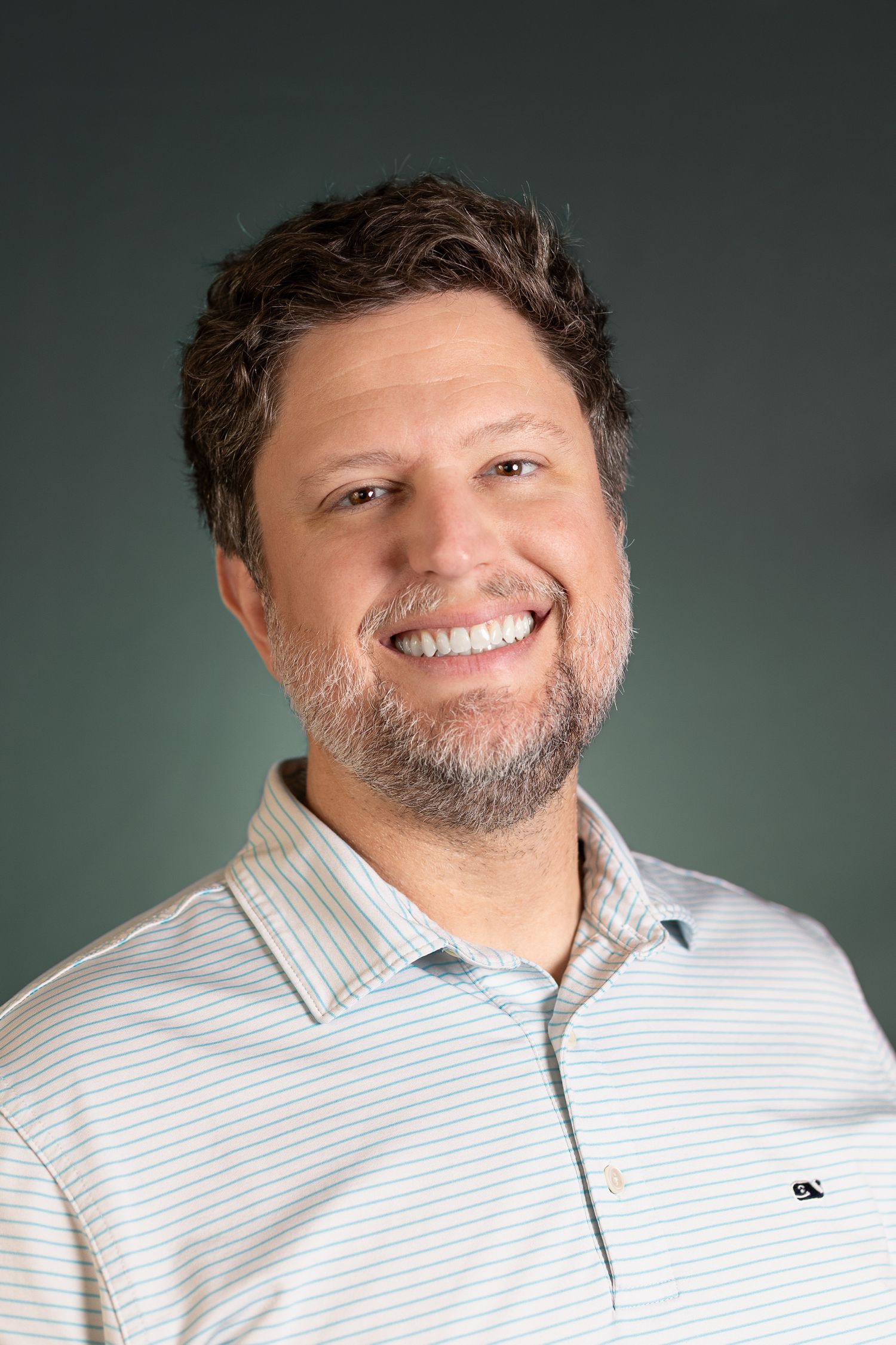 Professional headshot portrait of a person with a beard wearing a light striped collared shirt against a dark background.