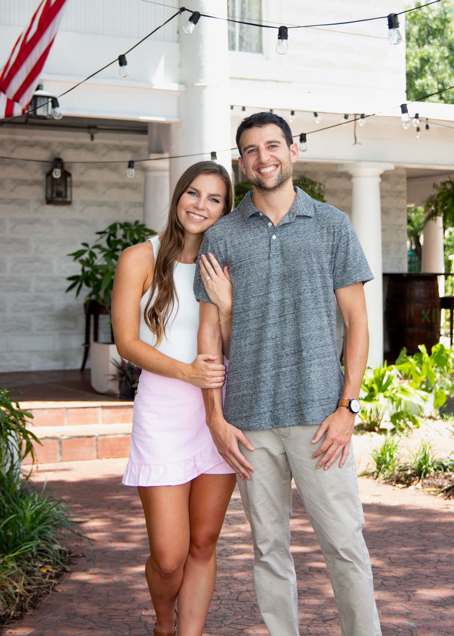A couple poses together on a brick patio with string lights and white columns visible in the background.