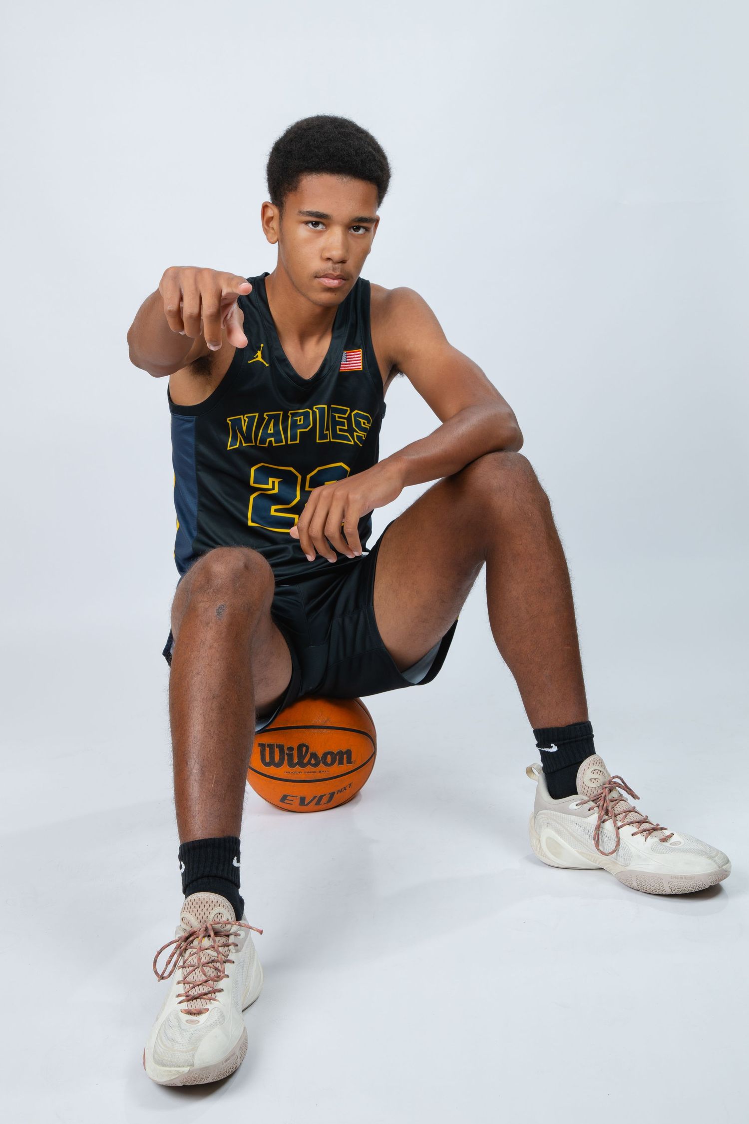 Basketball player in dark Naples jersey sits with Wilson basketball in studio setting against white background.