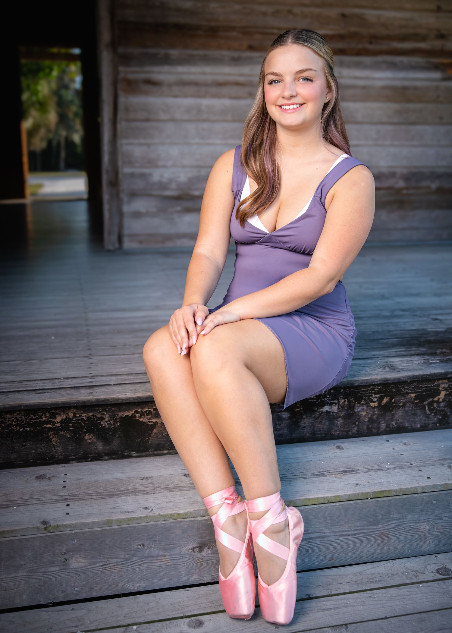 Ballet dancer in purple leotard poses with pink pointe shoes on rustic wooden porch.