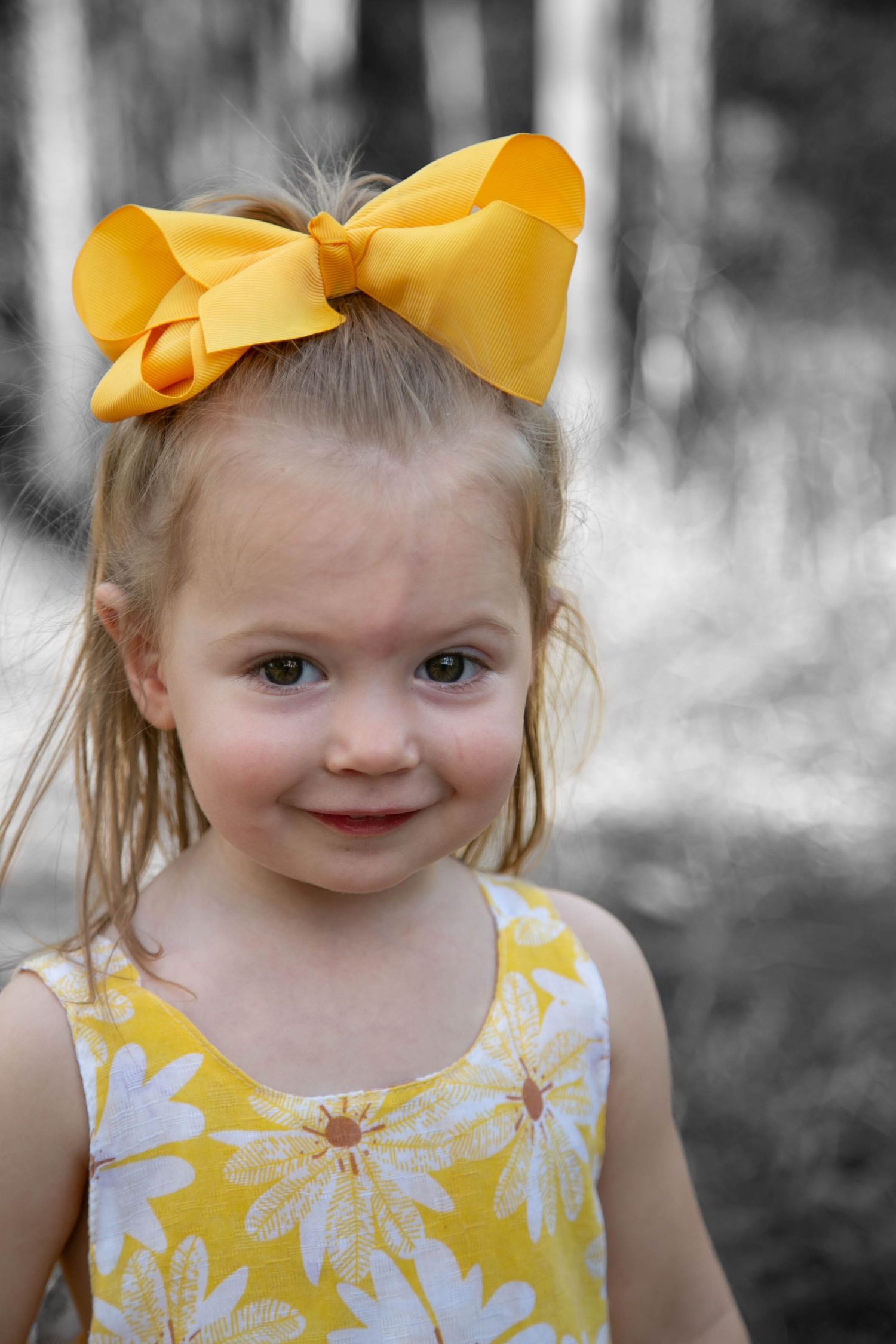 Young child wearing a yellow floral dress and large yellow bow against a black and white background.