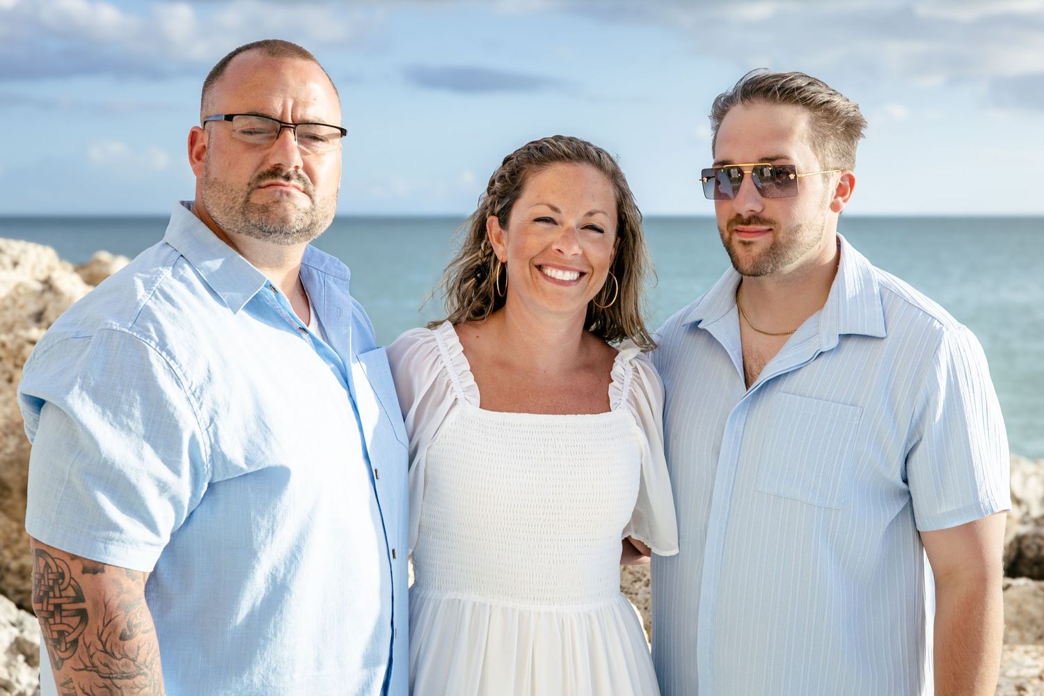 Three smiling people pose together on a rocky beach with blue ocean and sky behind them on a sunny day.