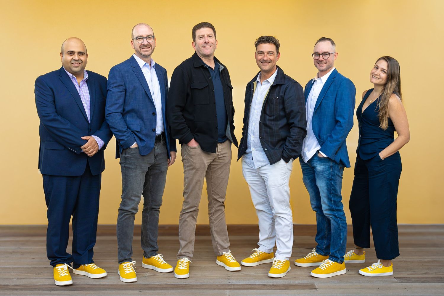 Six business professionals smiling and posing together in matching yellow sneakers against a bright yellow wall.
