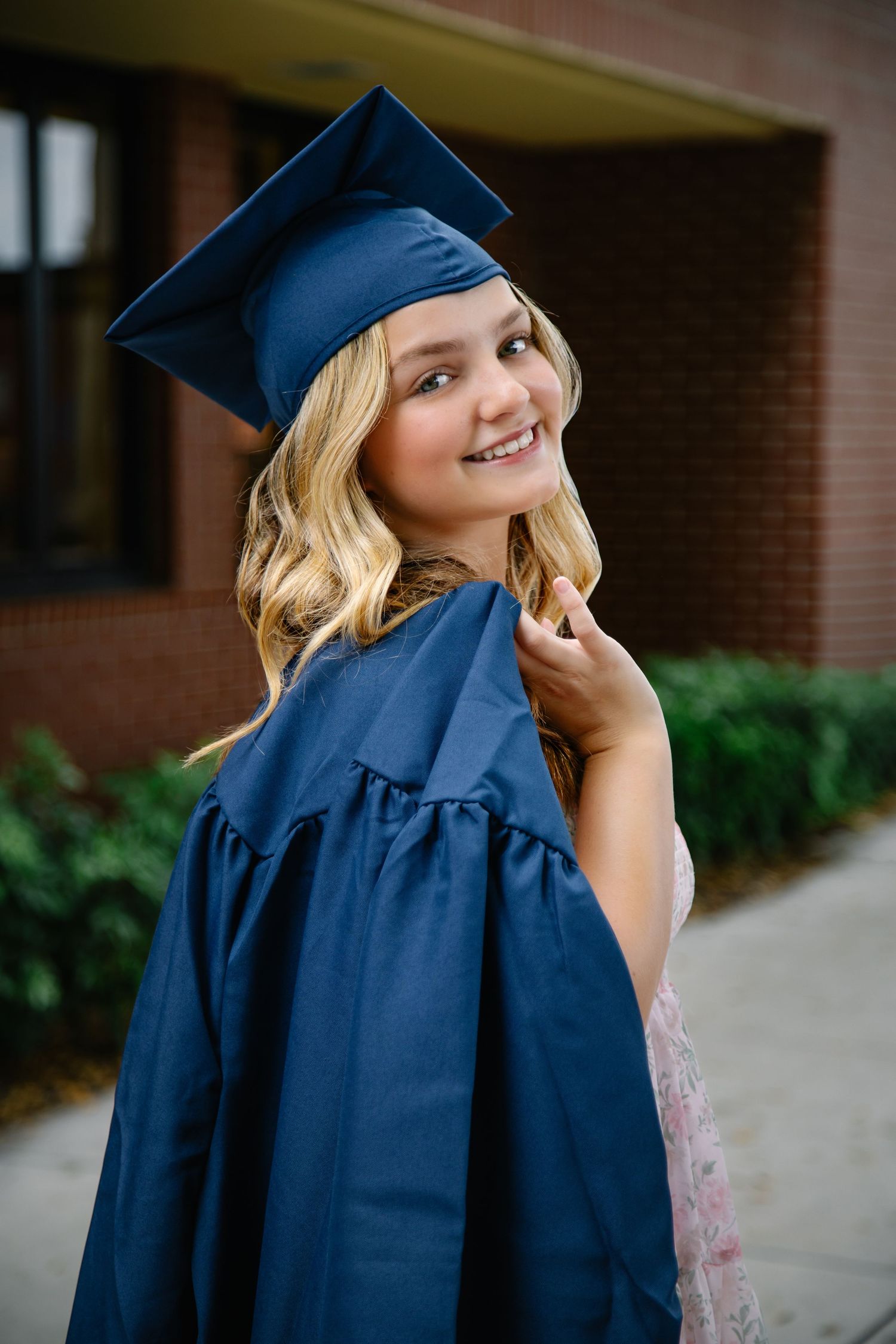 Graduate wearing navy blue cap and gown poses for portraits with a bright smile against brick building backdrop.