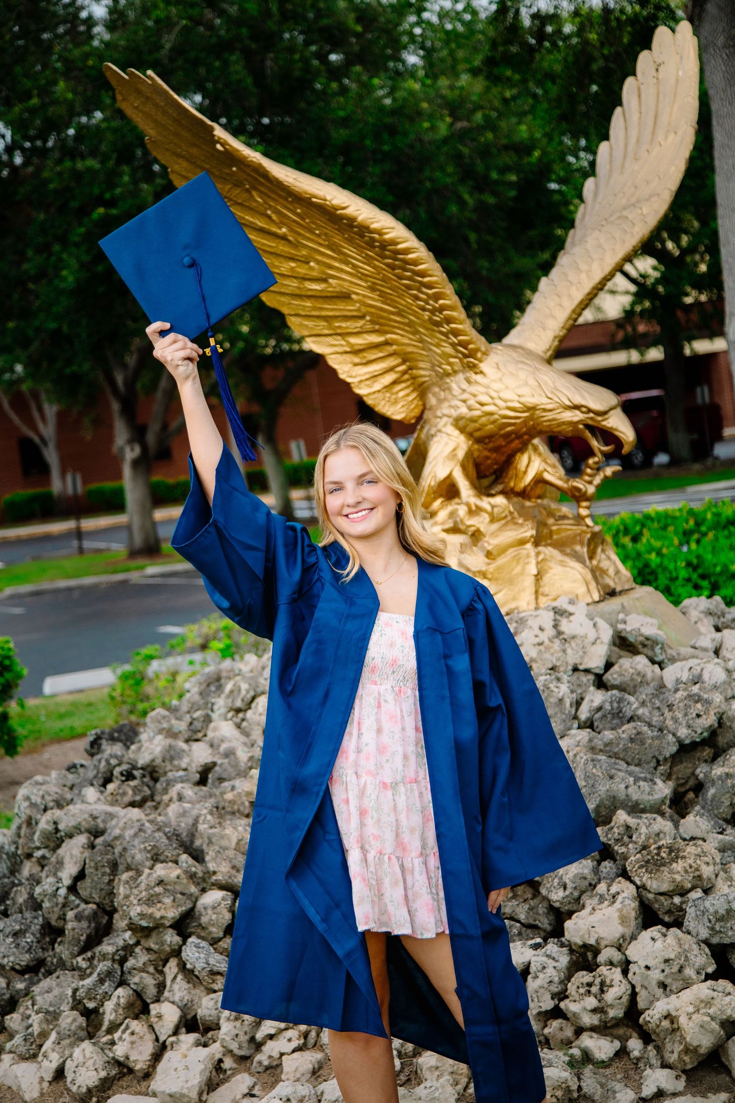 Graduate in blue cap and gown poses next to golden eagle statue during outdoor graduation photoshoot.