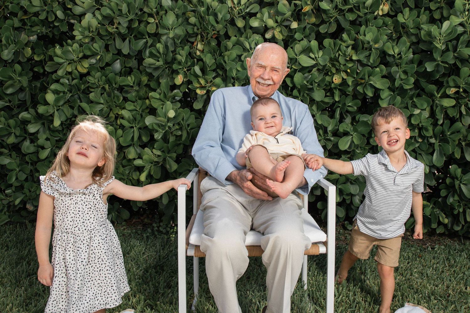 Four people sitting and standing together outdoors in front of a lush green hedge on a sunny day.
