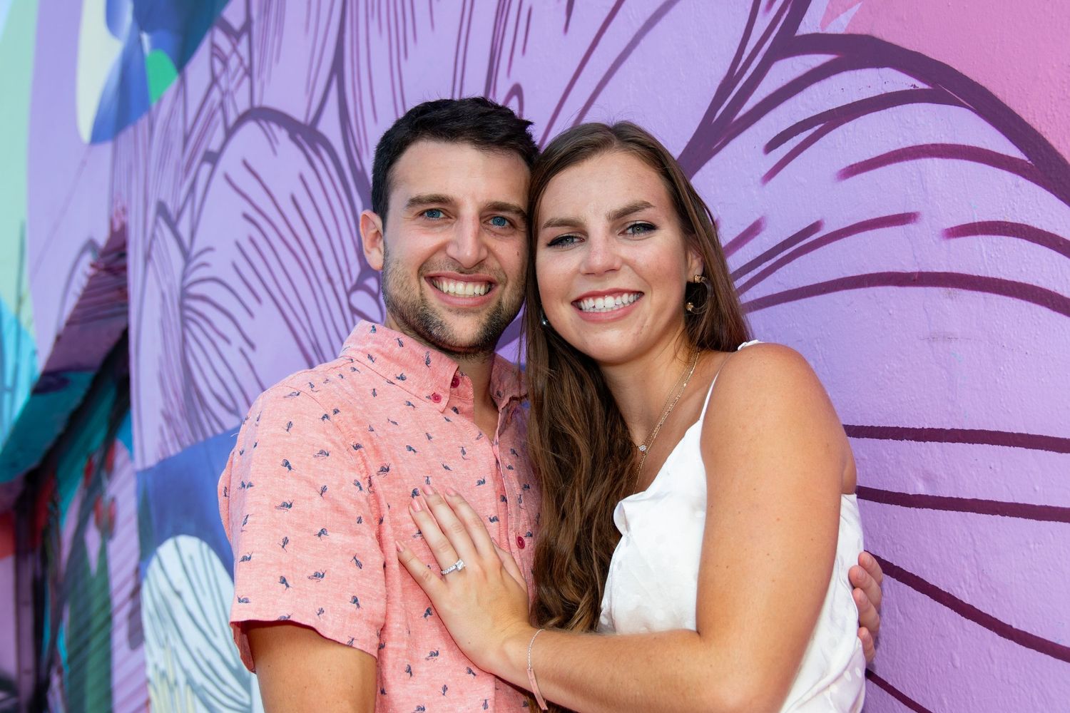 A couple shares a joyful moment together in front of a pink floral wall background.
