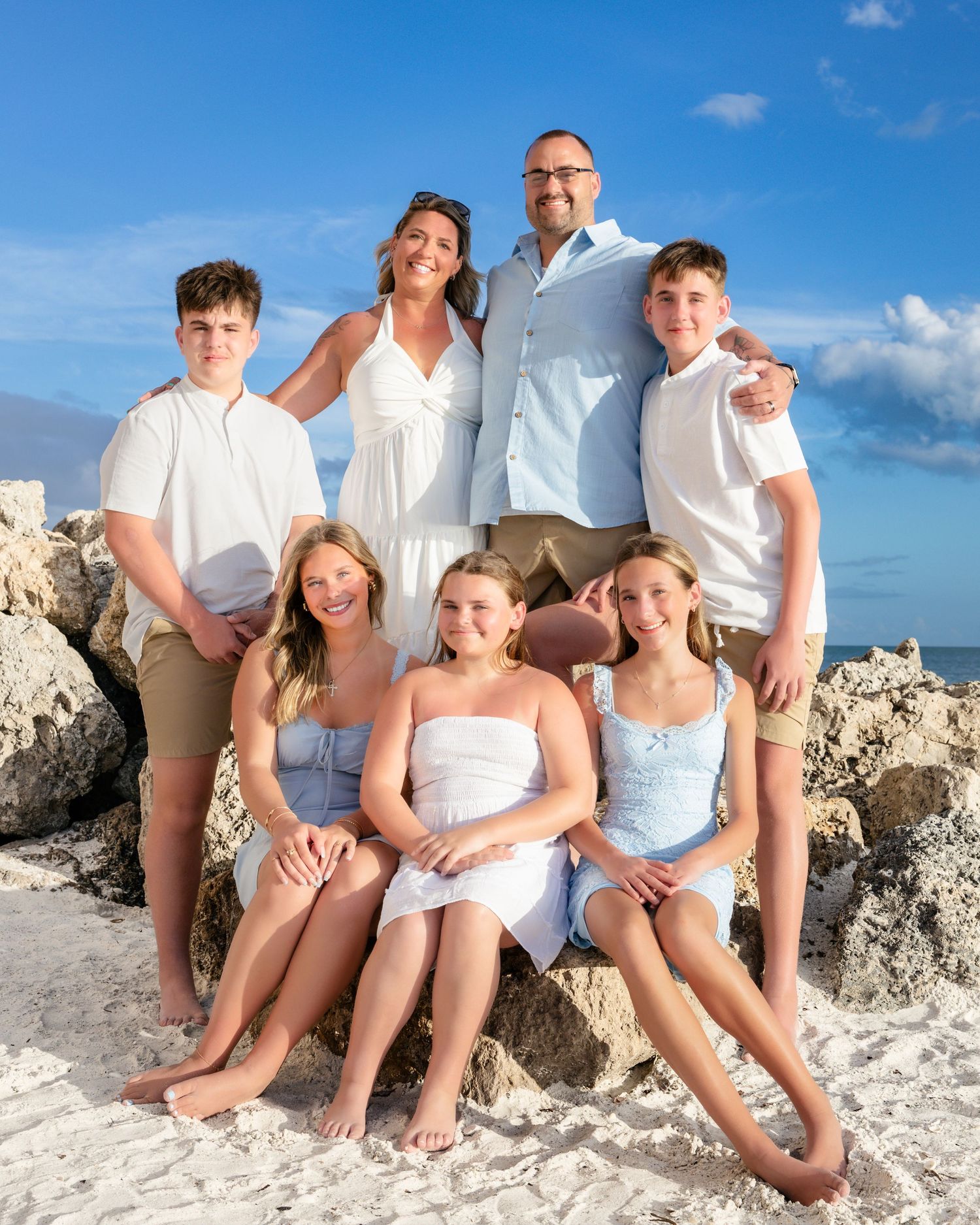 Happy family of seven posing on rocky beach with blue sky, wearing coordinated white and blue outfits.