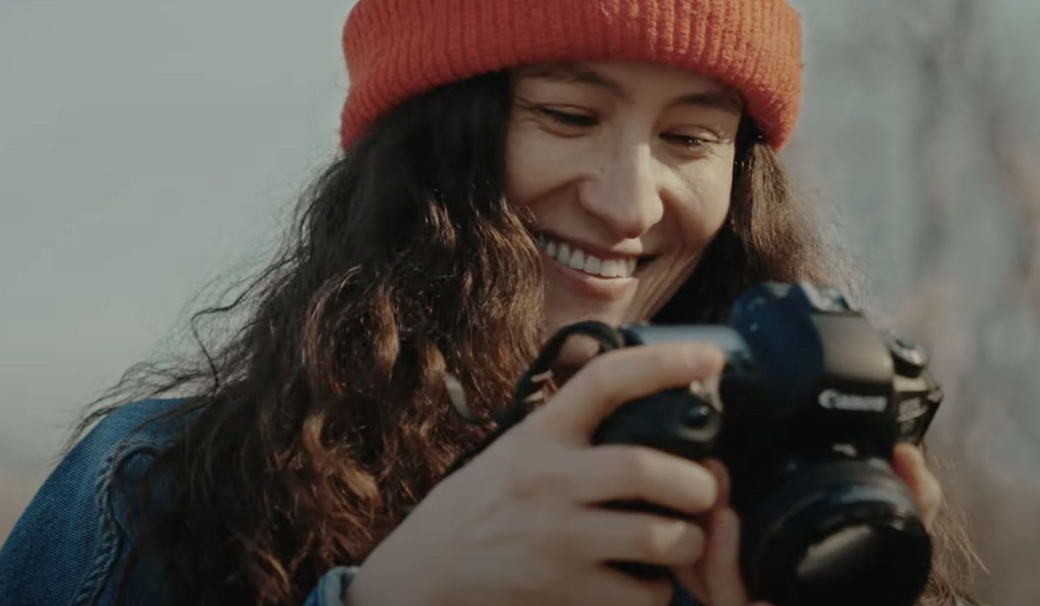 Smiling photographer wearing orange beanie and denim jacket reviews photos on digital camera outdoors.