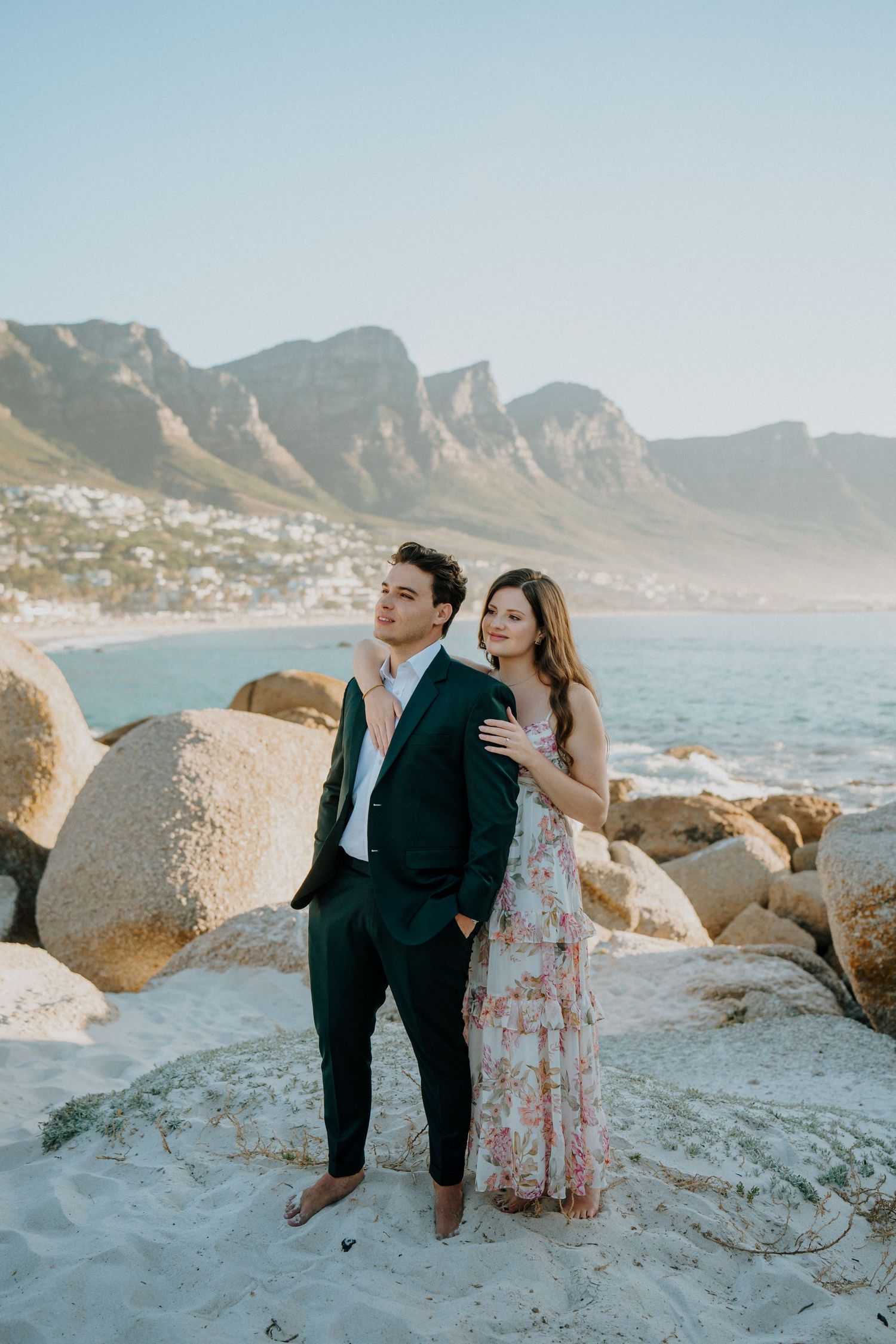 Couple poses together on rocky beach with dramatic mountain coastline and ocean views at sunset in Cape Town, South Africa.