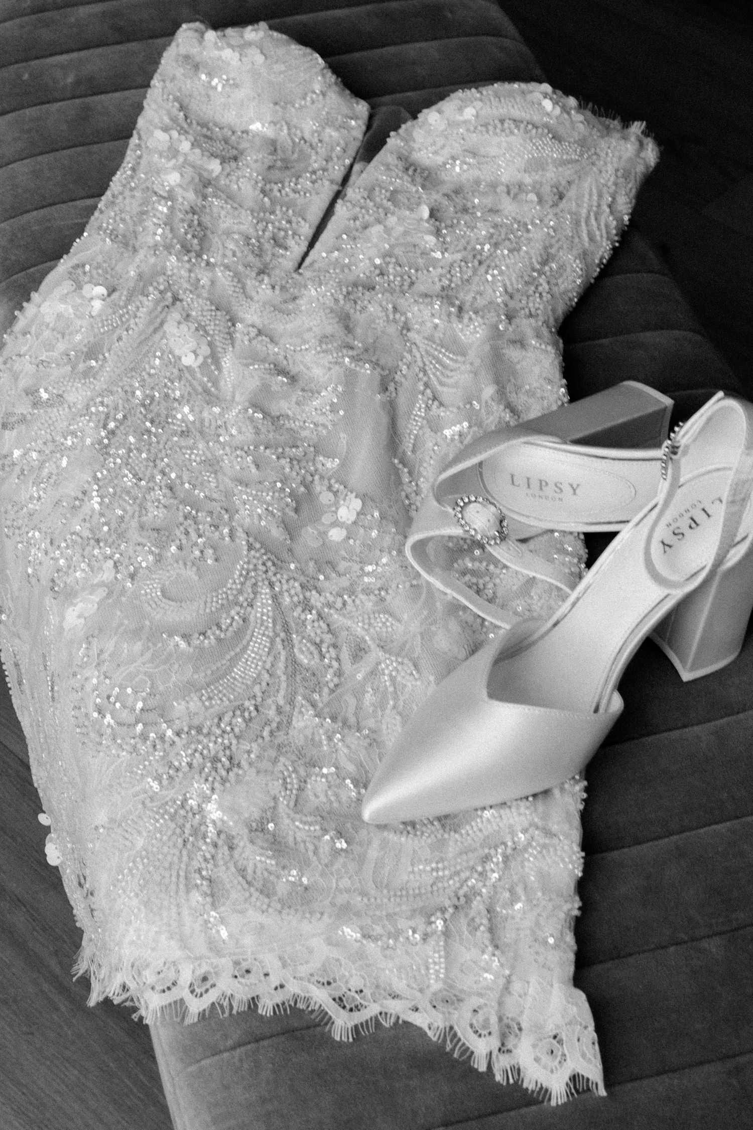 A detailed beaded white wedding dress bodice laid next to elegant silver bridal heels on a dark wooden floor.