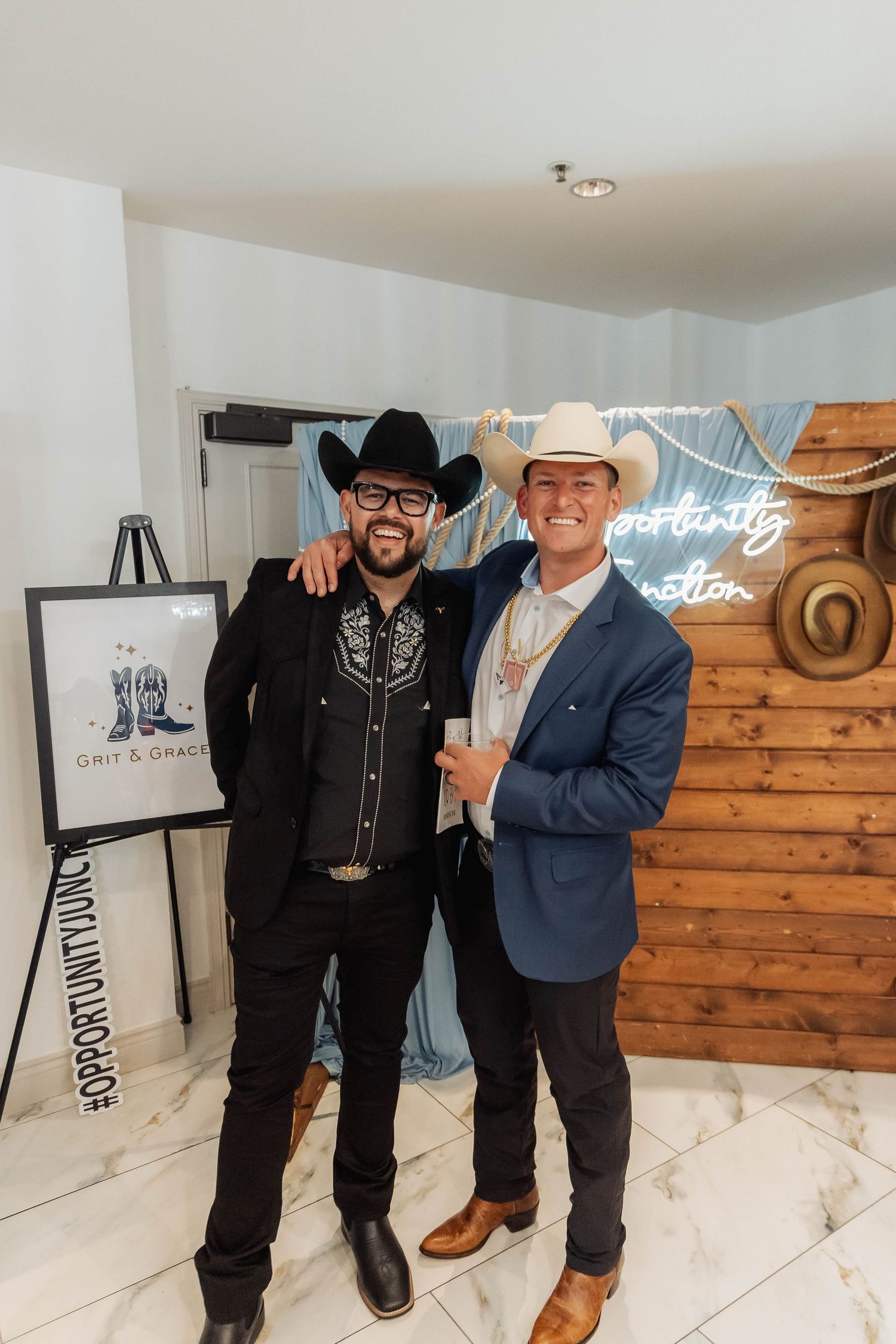 Two men in cowboy hats and western attire pose at a Grit & Grace event near a rustic wood wall.