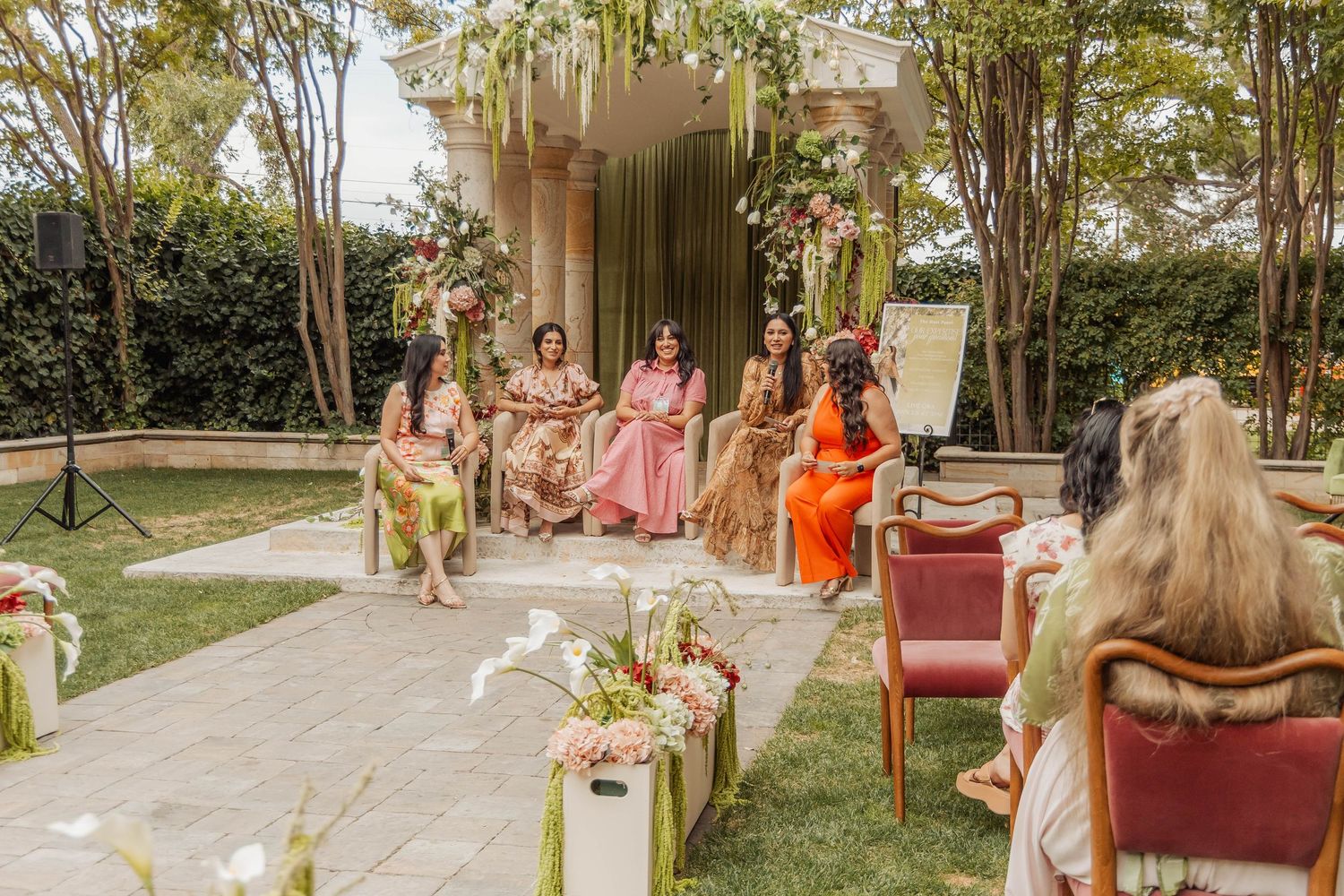 Women in colorful traditional attire seated outdoors at an elegant floral-decorated Indian wedding ceremony.