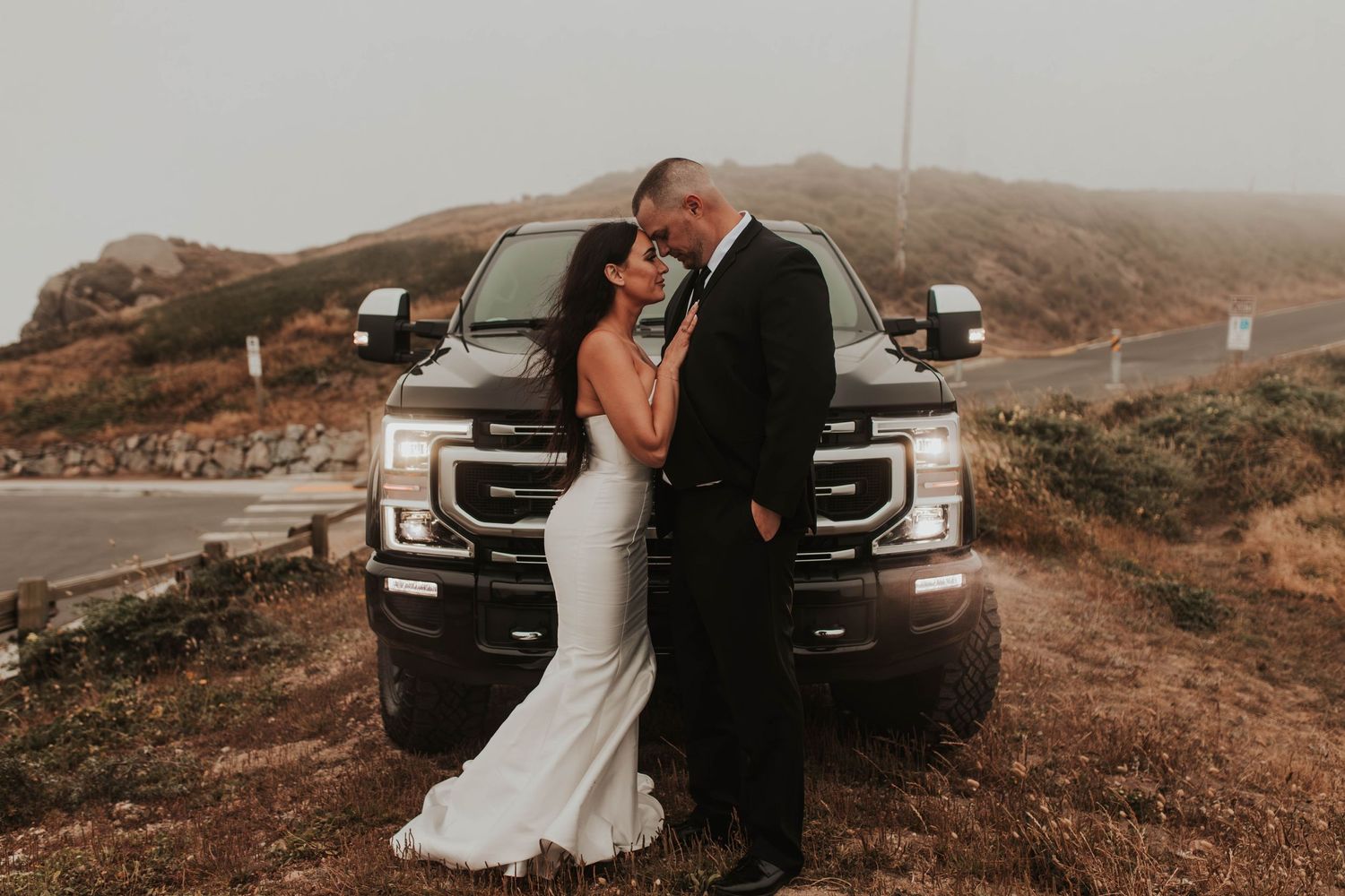 A wedding couple poses in front of a large pickup truck on a foggy mountain road.