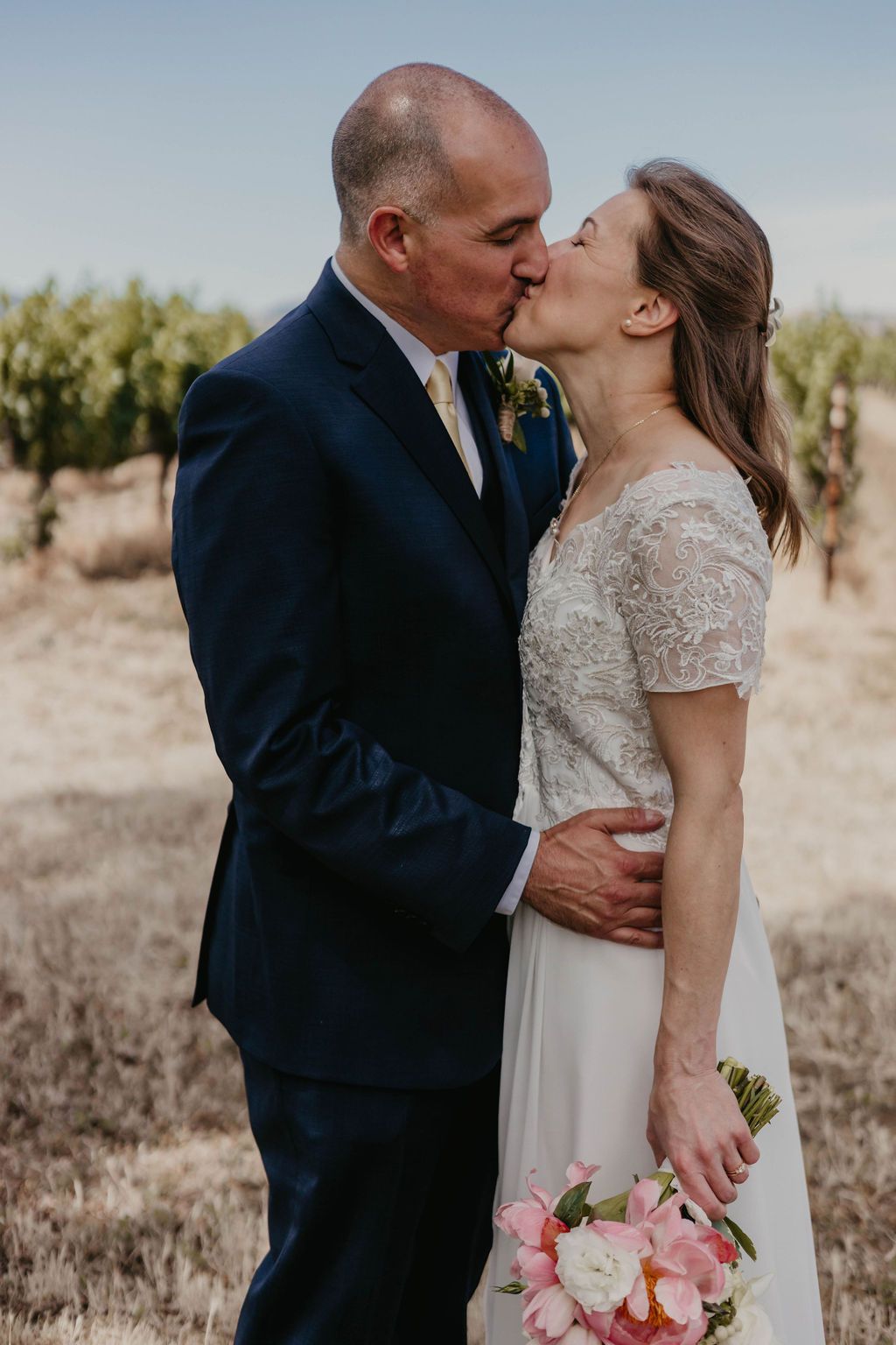 Intimate kiss shared between newlyweds in rustic vineyard setting with pink bouquet.