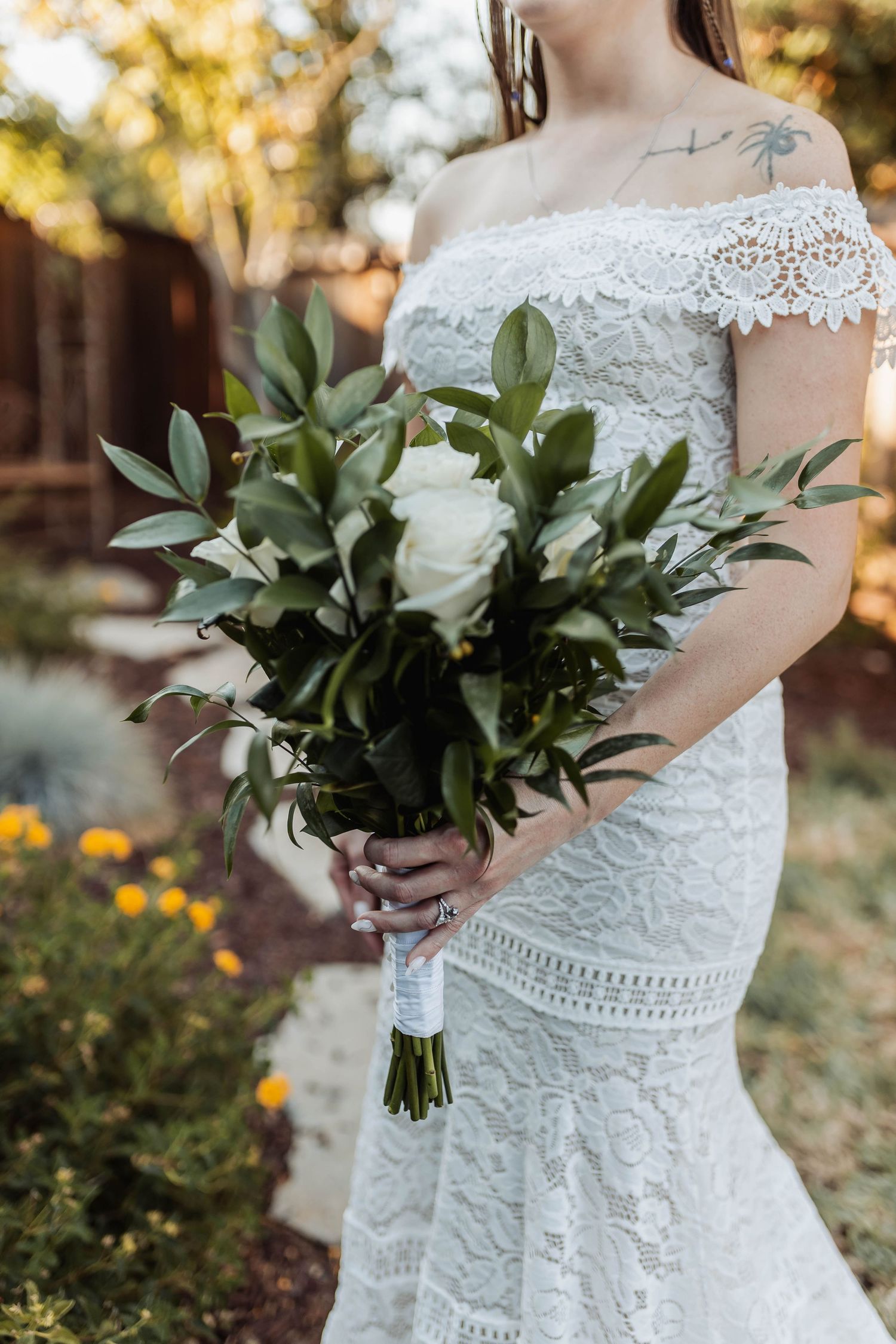 A bride in lace dress holds a classic white and green wedding bouquet in an outdoor garden setting.