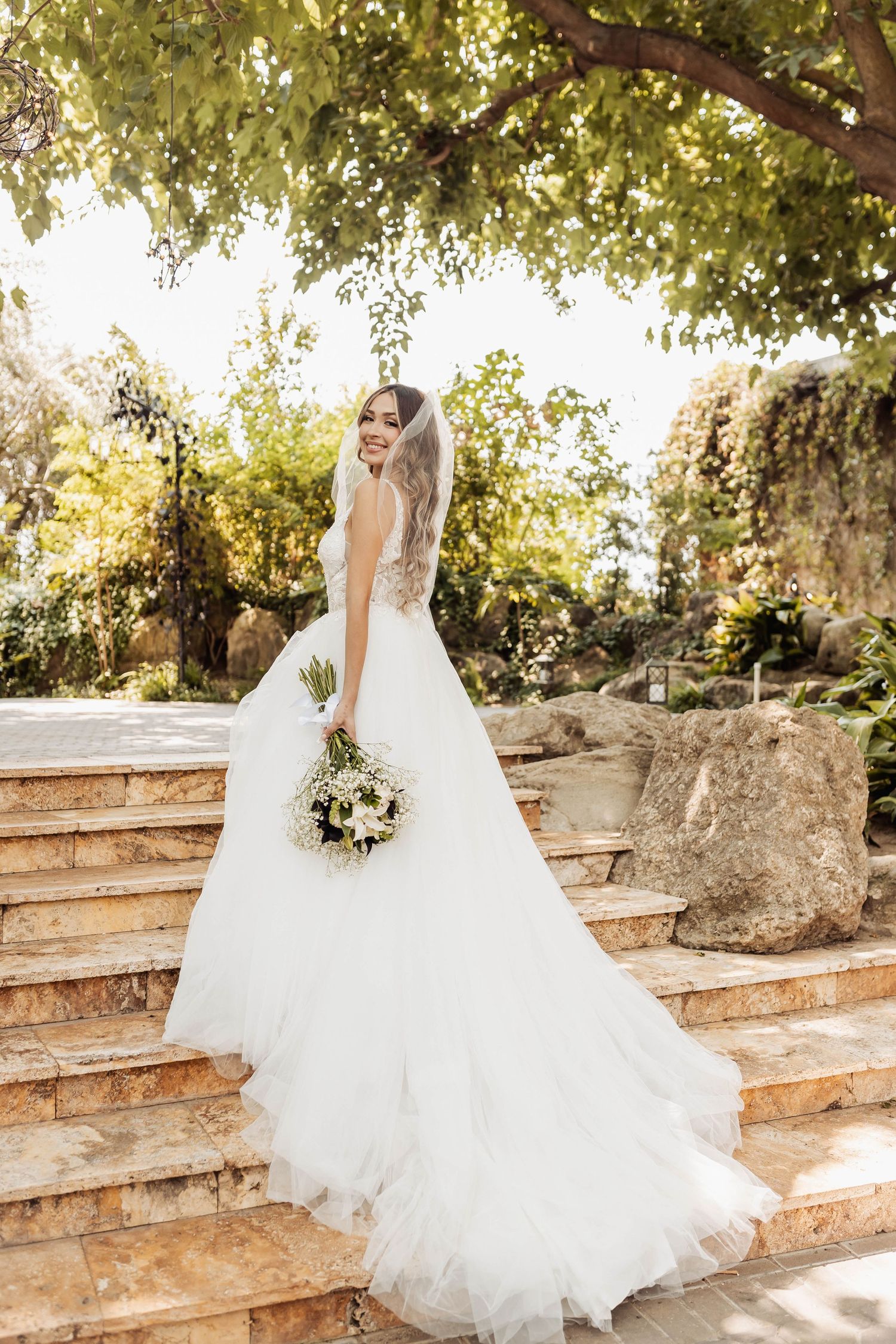 Elegant bridal portrait on rustic stone steps with white bouquet and flowing dress.