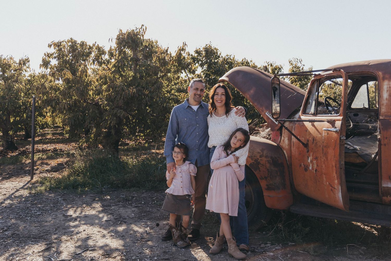 Family of four posing beside a rustic vintage red truck in an orchard setting on a sunny day.