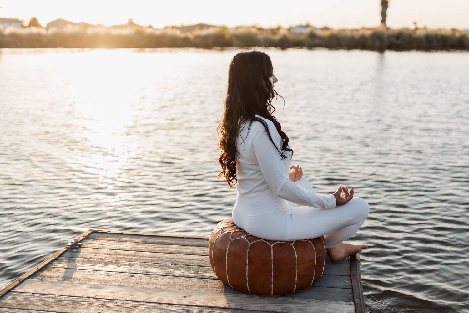 Person meditating on a leather pouf on a wooden dock at sunset.