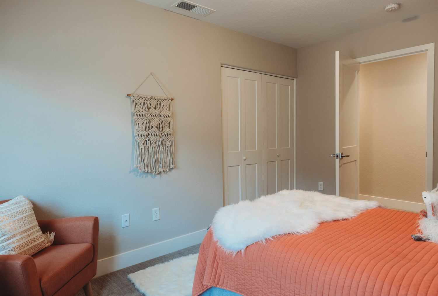 Minimalist bedroom with coral bedspread, macrame wall hanging, and comfortable armchair.