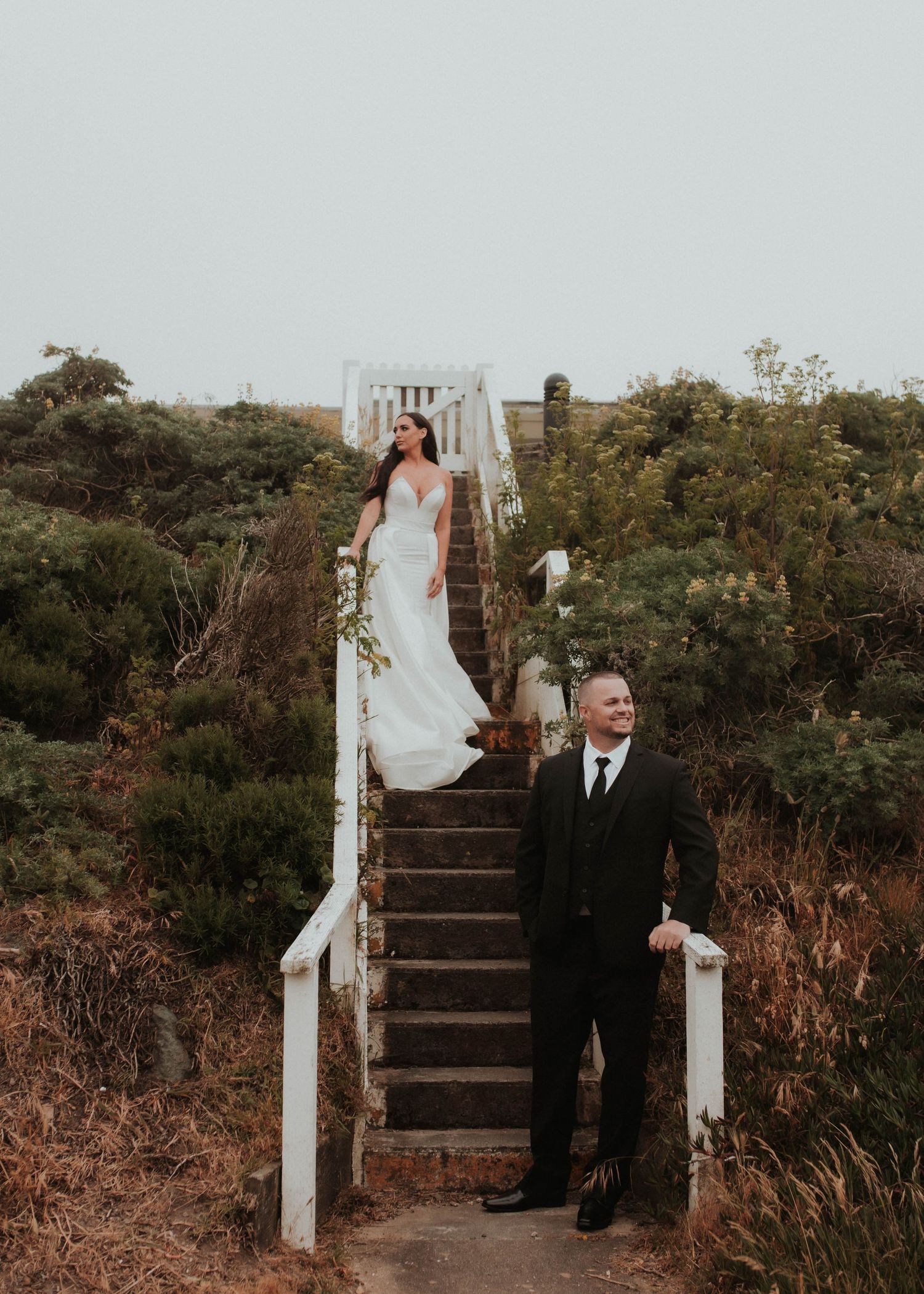 Wedding photo on rustic white stairs with bride above and groom below amidst coastal vegetation.