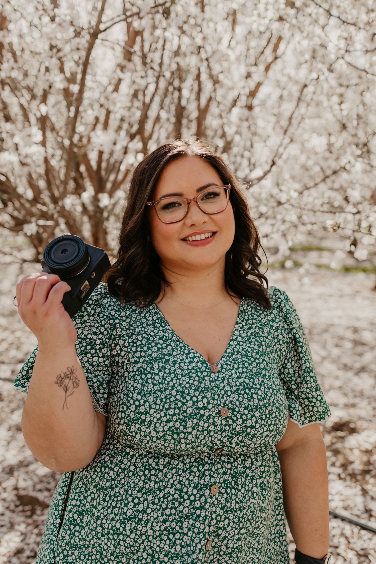 Professional photographer with camera poses in front of blooming cherry blossoms wearing a green floral print dress.