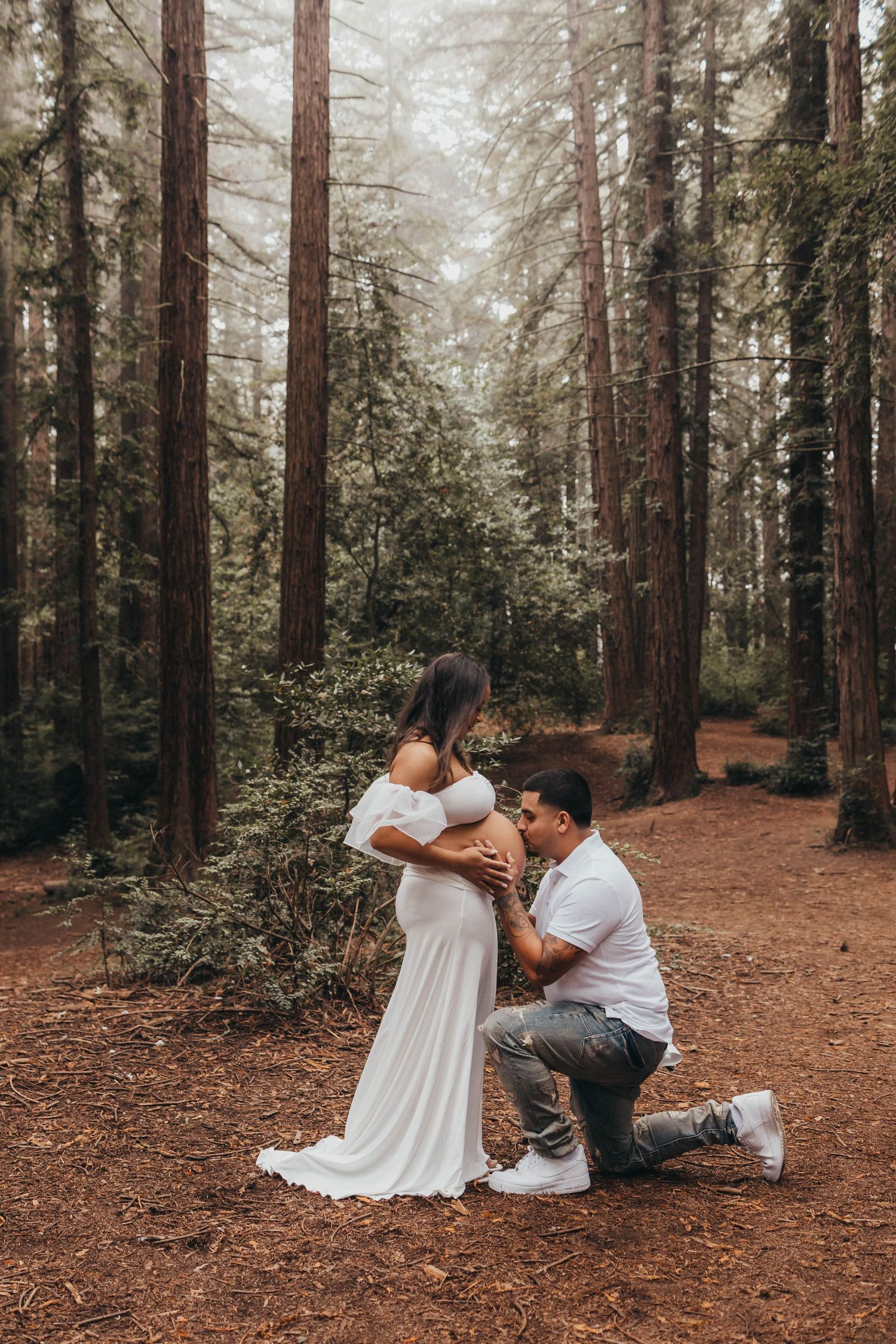 Man kneeling before pregnant woman in white gown among tall redwood trees in a romantic maternity photo shoot.