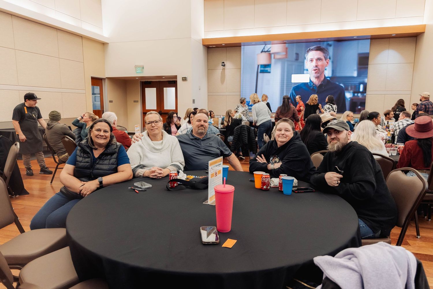 Group of people smiling at a round table during a community event in a large hall with a presentation screen behind them.