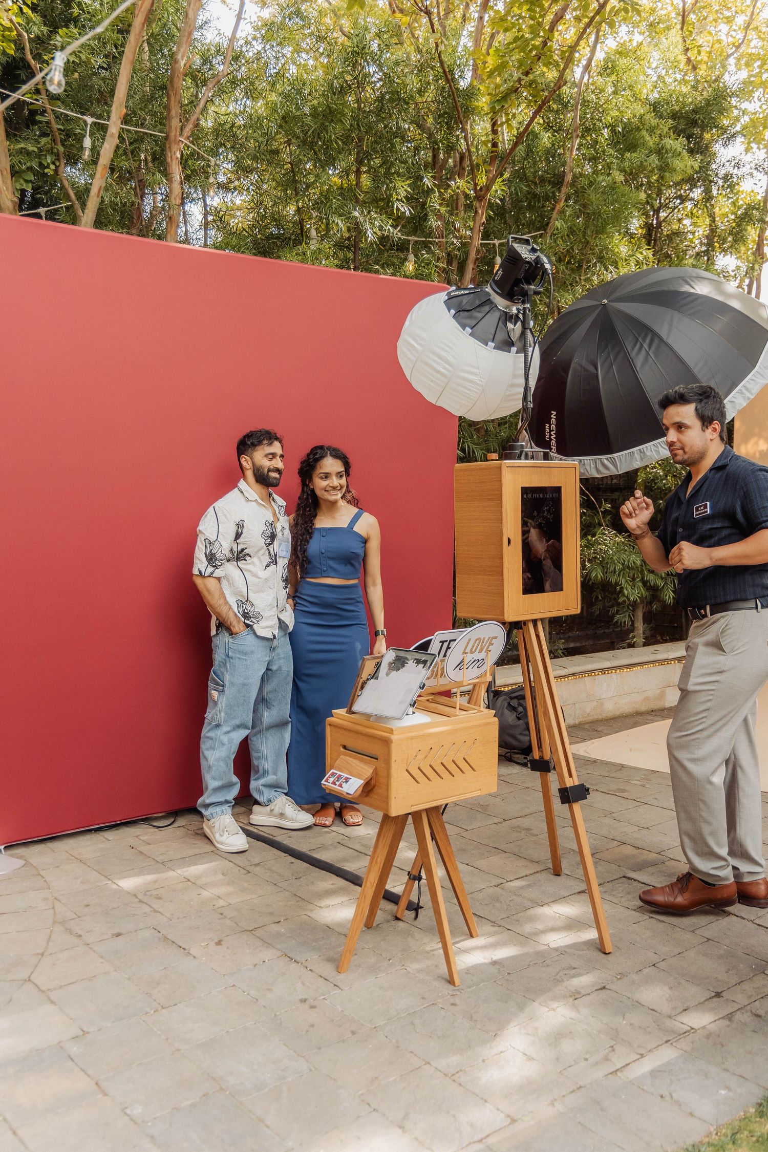 Couple posing in front of red backdrop at outdoor photo shoot with professional lighting equipment and photographer.