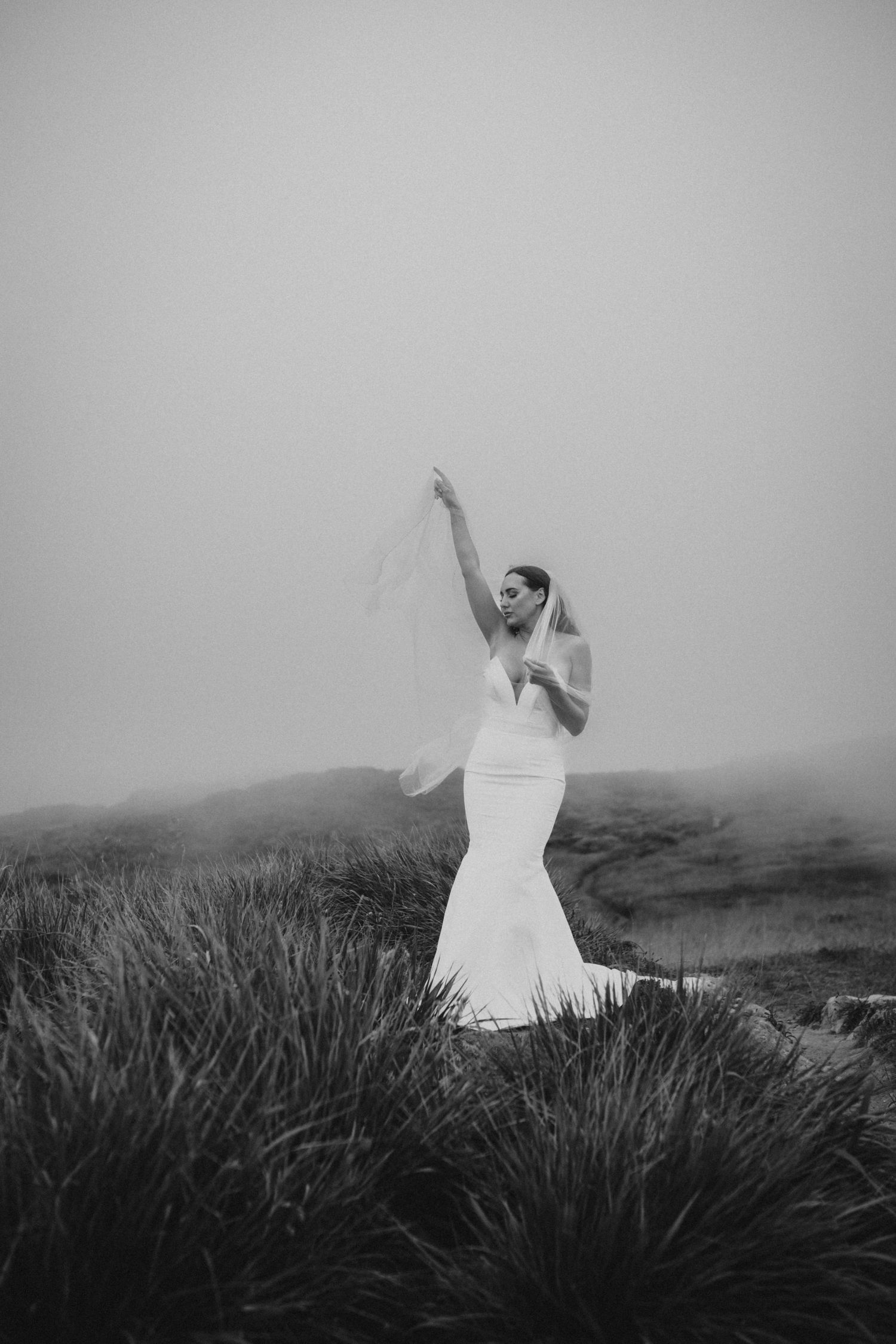 Black and white photo of a bride in a mermaid dress posing on a grassy hill in misty conditions.