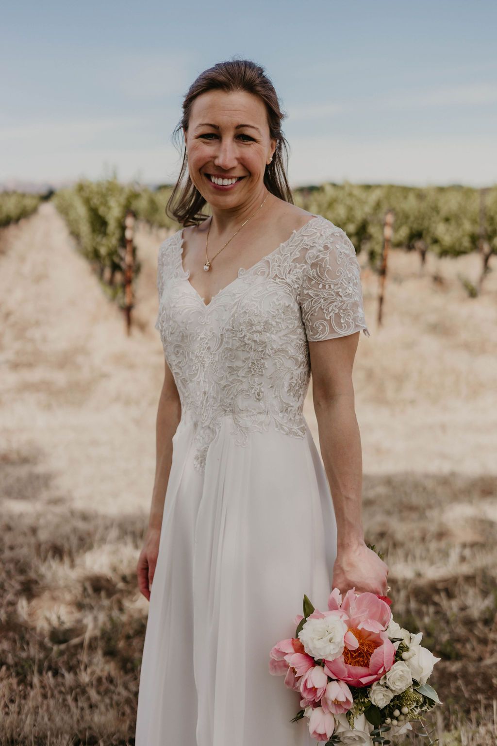 Joyful bride in lace-detailed wedding dress poses in vineyard setting with pink bouquet.