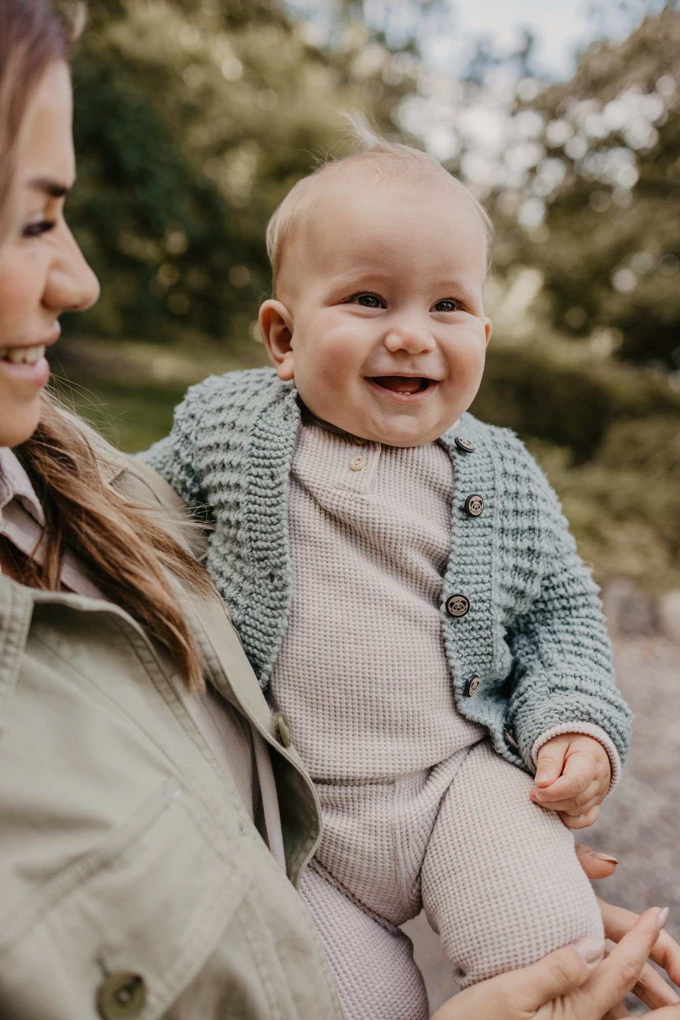 Close-up of smiling baby wearing gray knit cardigan held in loving embrace outdoors.