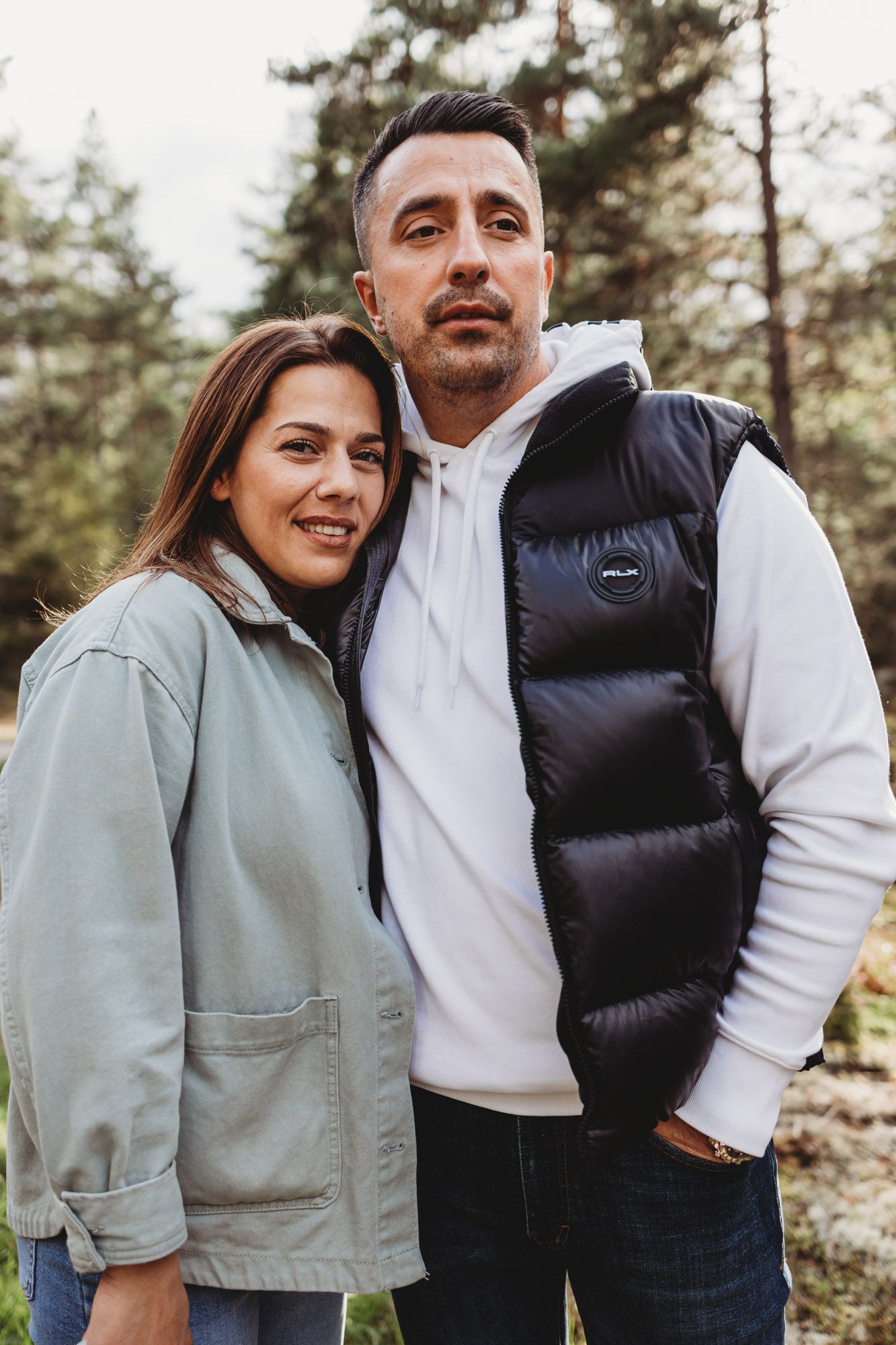 A casual outdoor portrait of a couple in matching puffer vests against a pine forest backdrop.