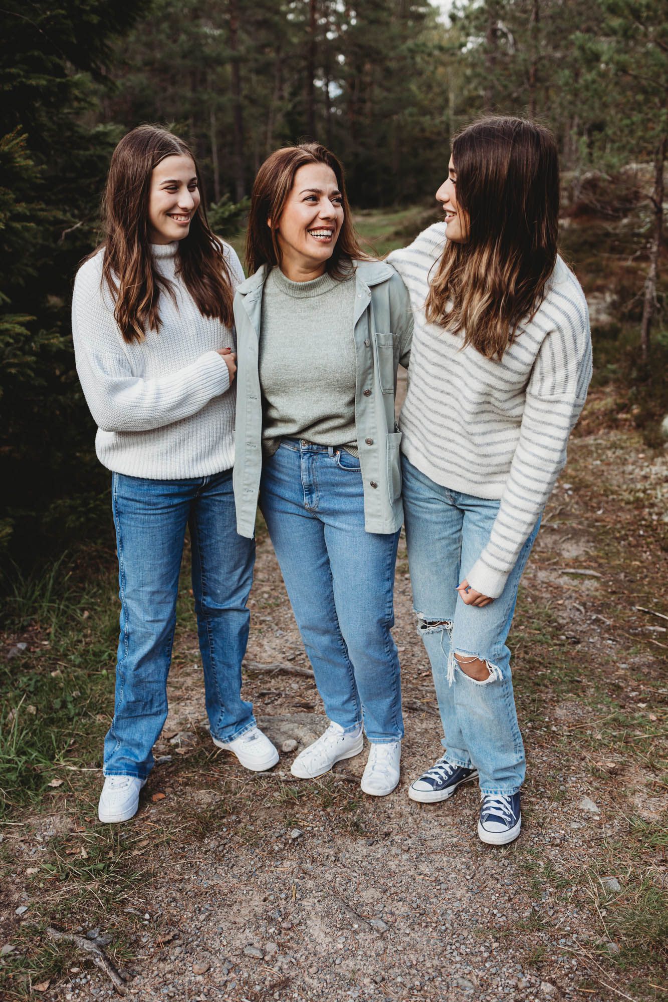 Three friends in cozy sweaters and jeans share laughter while standing on a forest path.