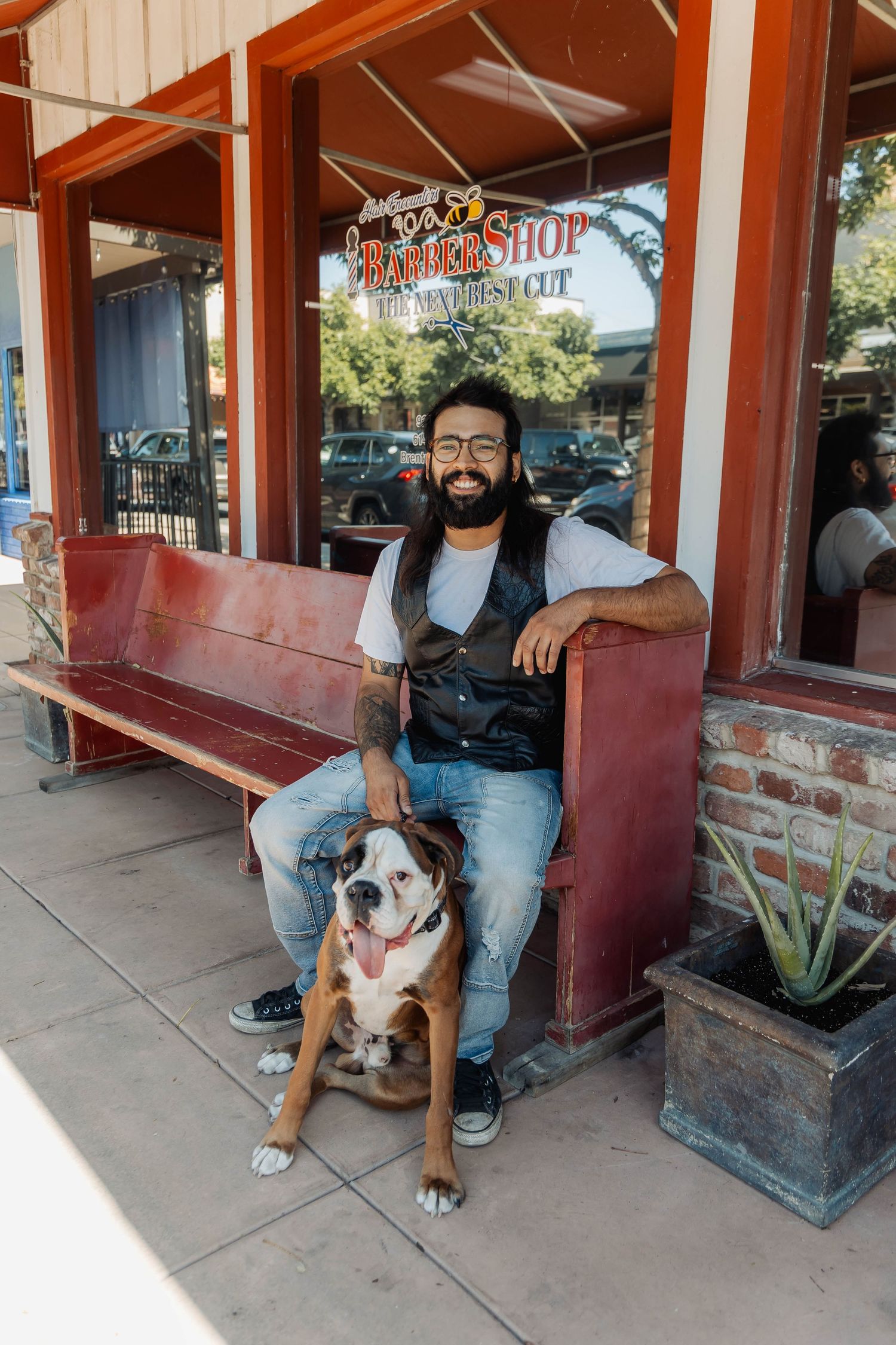 Casual portrait of a person with their dog sitting on a red bench outside a barber shop.