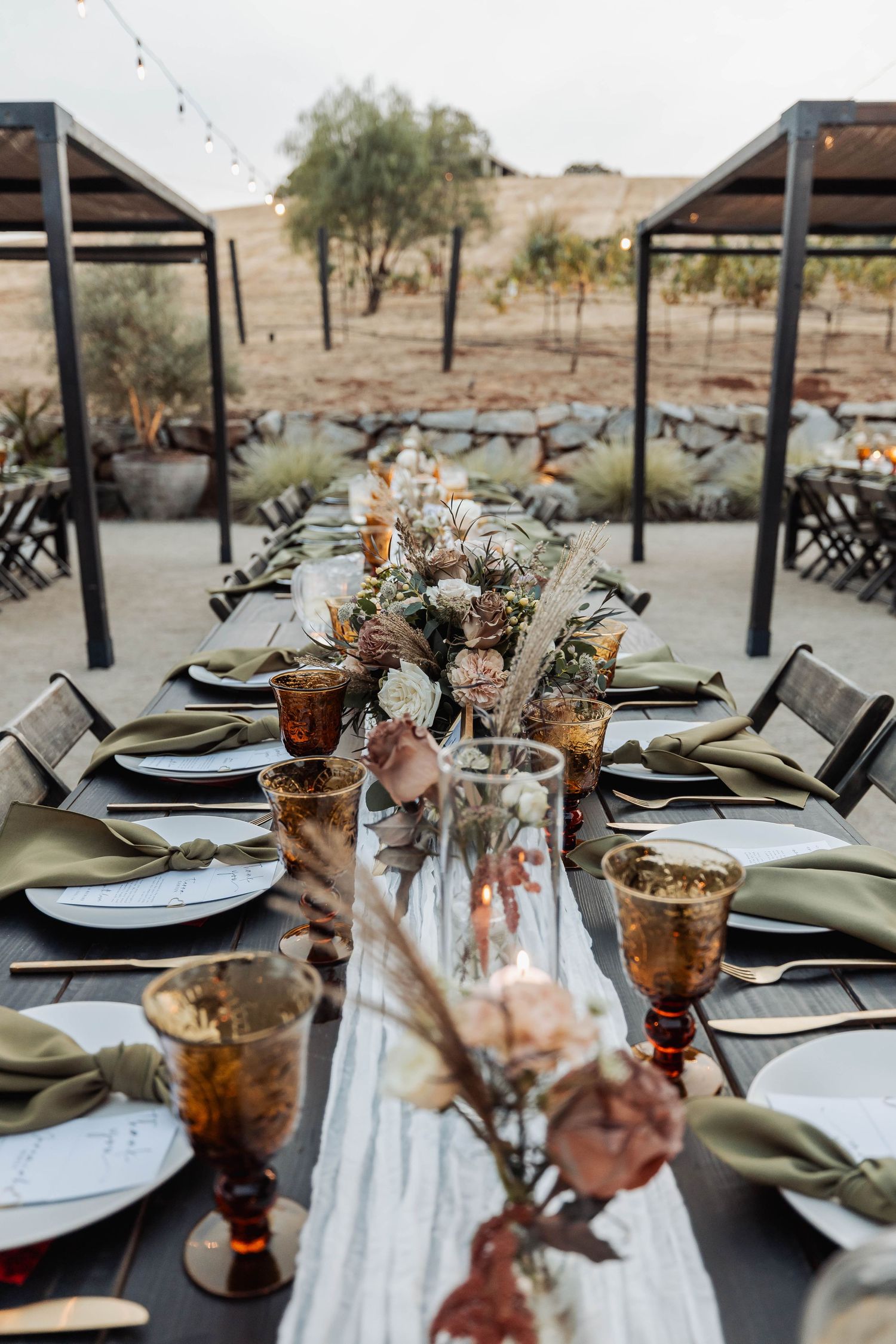 Rustic outdoor wedding reception table settings with copper glasses and sage green linens in a desert landscape.