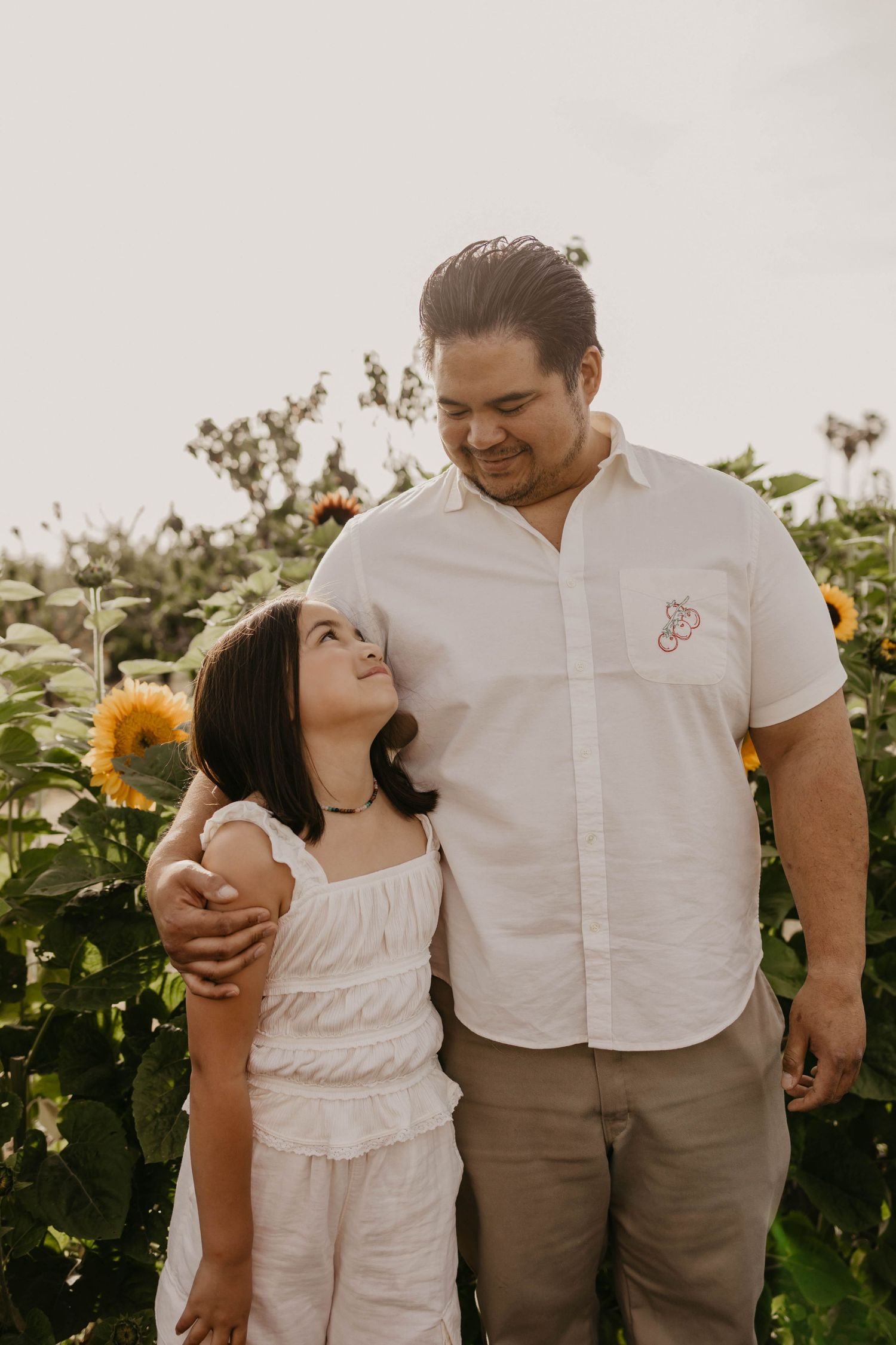 Two people sharing a tender moment in a sunflower field at sunset, with warm golden light and natural surroundings.