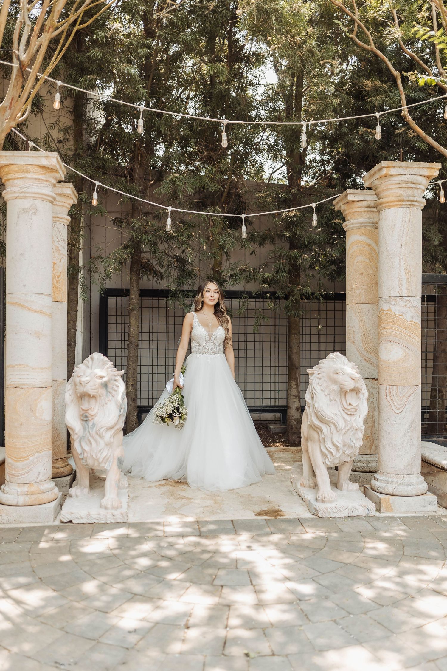 Wedding dress portrait between classical stone columns with decorative lion statues and string lights.