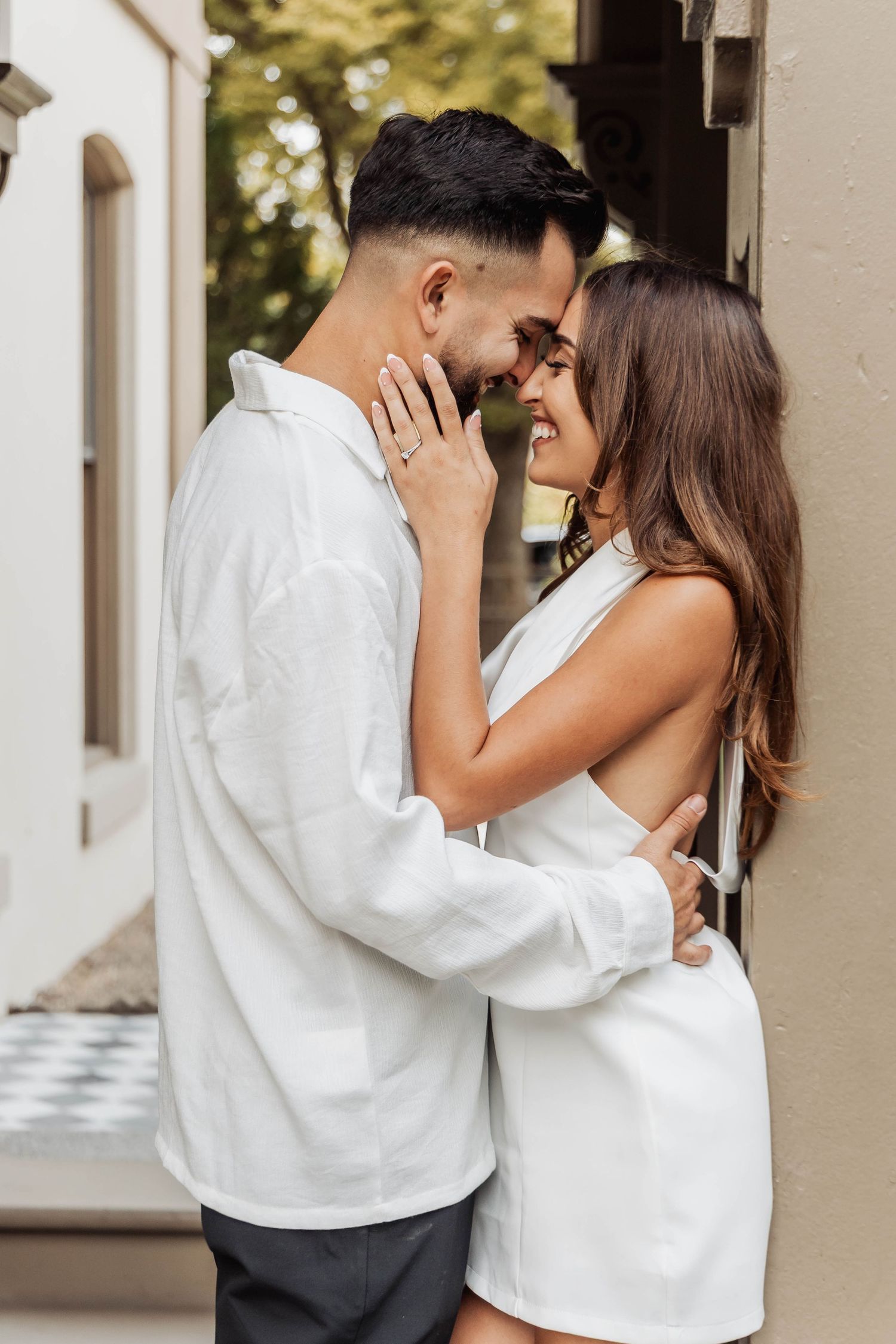 Romantic couple in white outfits sharing tender moment against architectural backdrop.