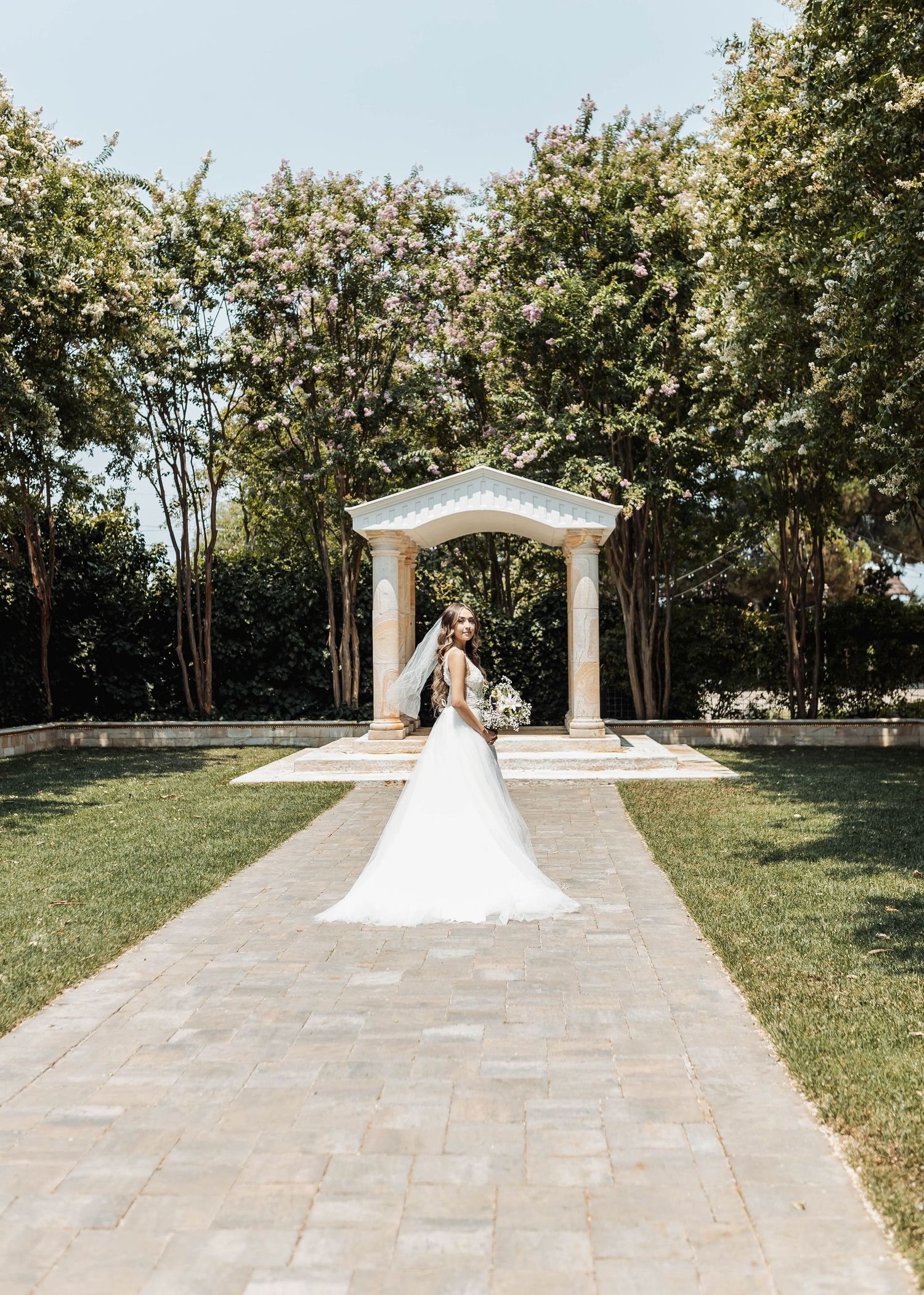 Wedding dress photographed in front of white classical garden arch with manicured landscaping.