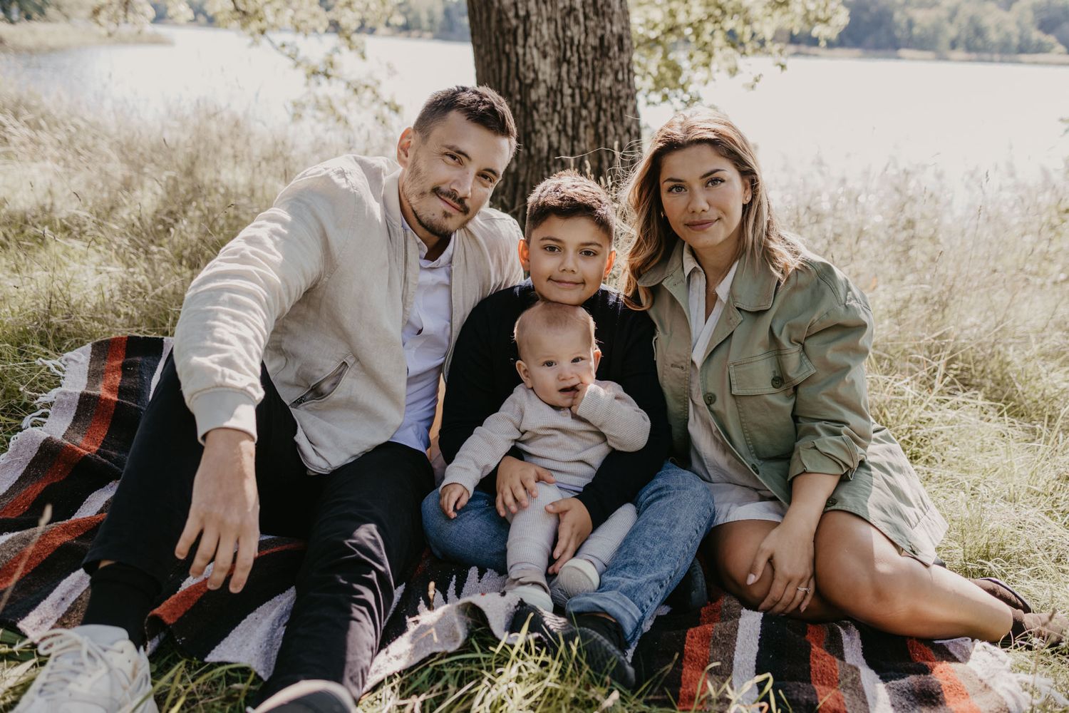 A family of four sits together on a plaid blanket outdoors on a sunny autumn day.