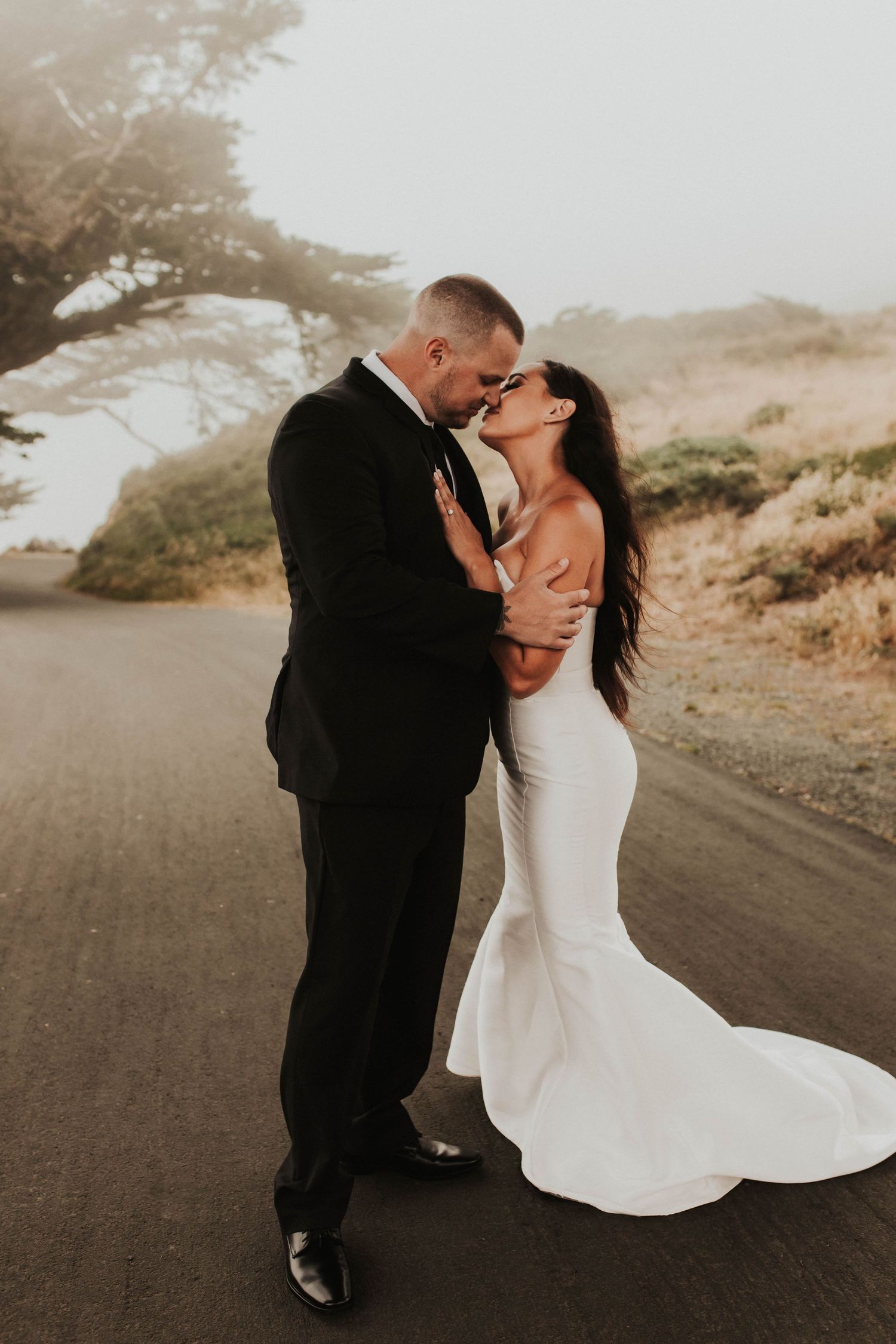 Newlyweds share an intimate moment on a misty mountain road at sunset.