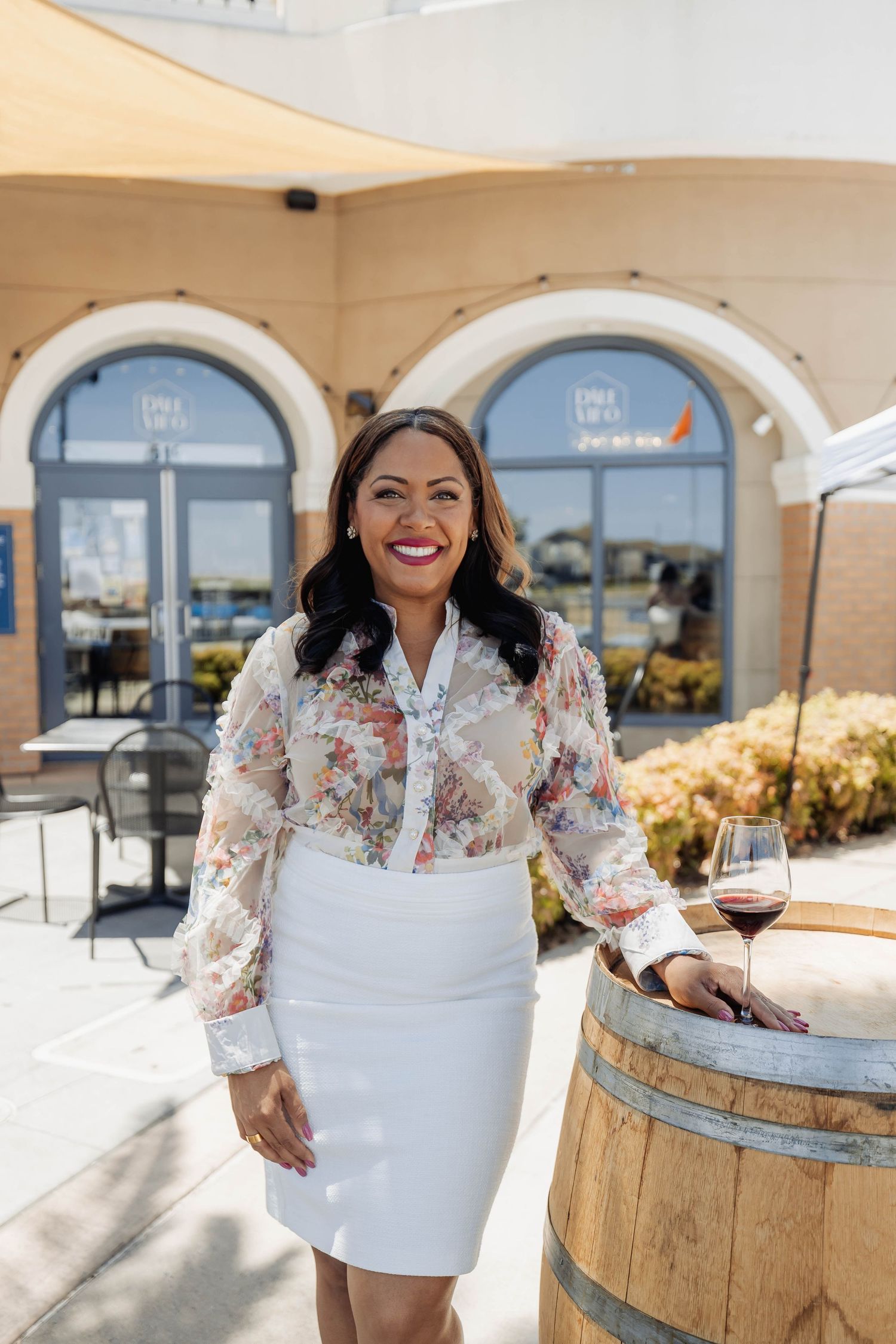 Individual in floral blouse poses near wine barrel on outdoor patio.