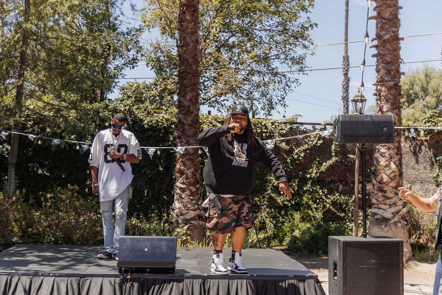 Performers on outdoor stage at Raiders themed fan event with speakers and palm trees background.