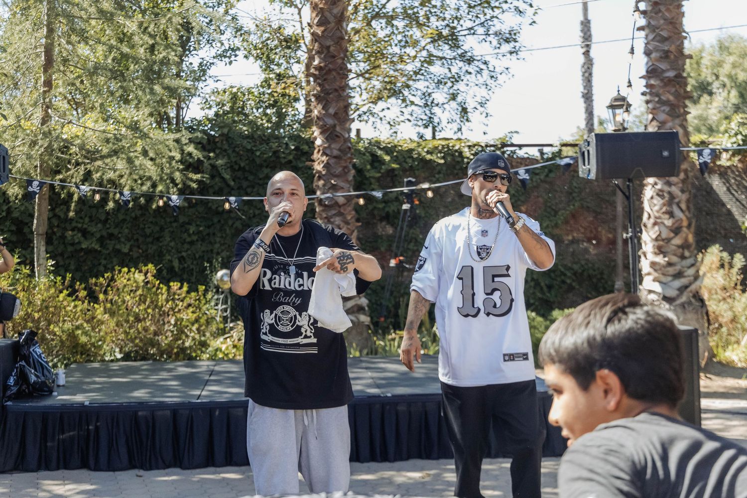 Two men performing on outdoor stage, one in Raiders shirt, one in jersey number 15, with audience watching.