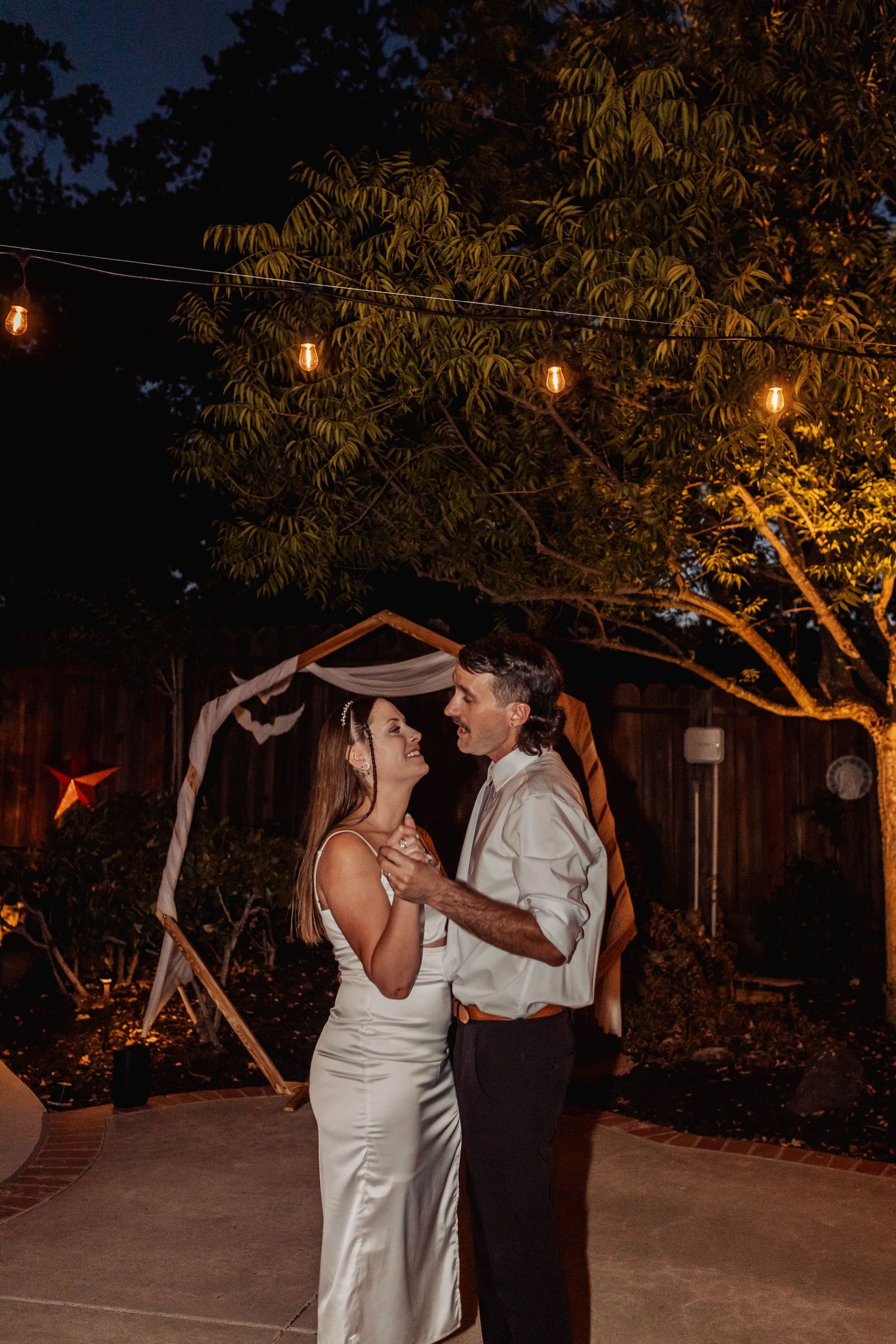 Newlyweds dancing under string lights at an outdoor evening wedding reception.