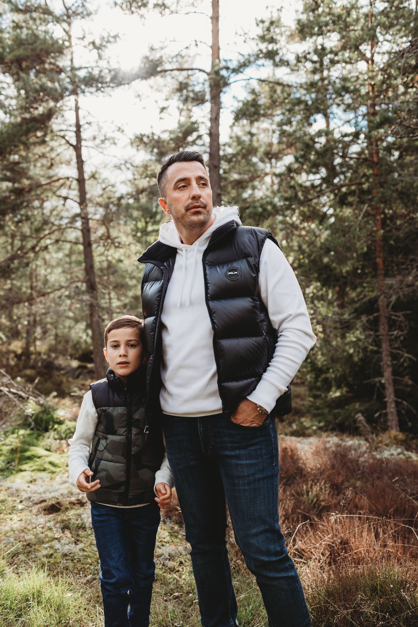 A matching pair in puffer vests and white sweaters stand together in a wooded setting.