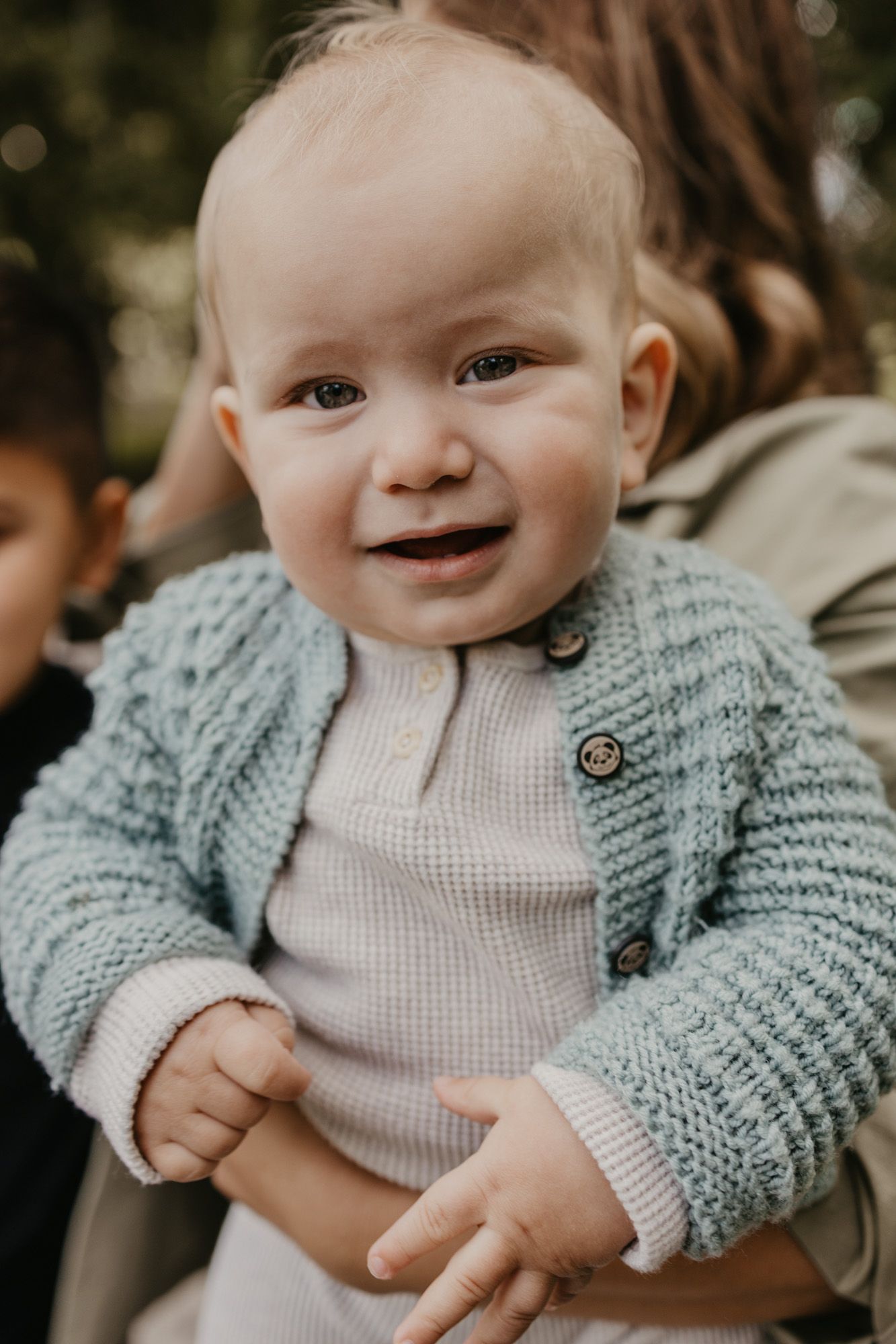 Baby with bright expression wearing cozy gray cardigan in outdoor setting.