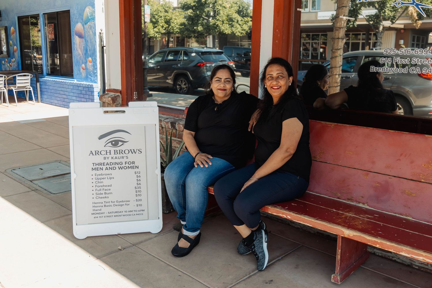 Two people sitting together on a red bench outside a barber shop with a sidewalk sign.